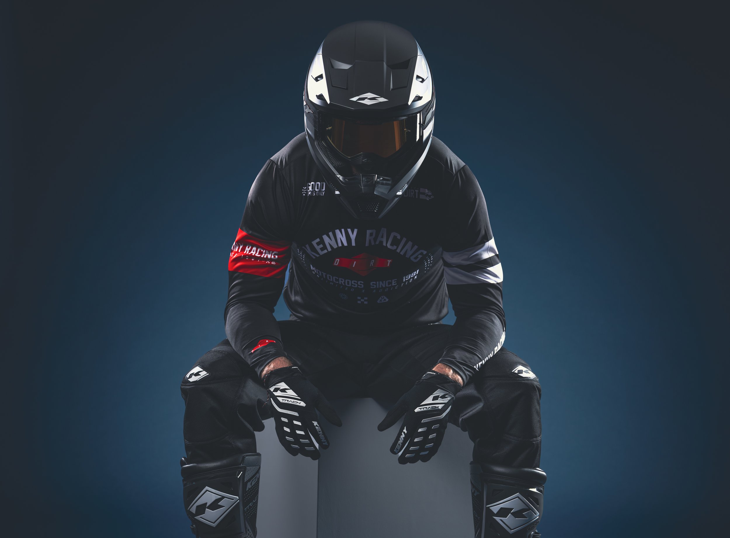 Un pilote de motocross en position centrée, portant un casque noir, une combinaison noire avec des détails colorés, et des gants et bottes de motocross de style sportif, sur fond sombre.