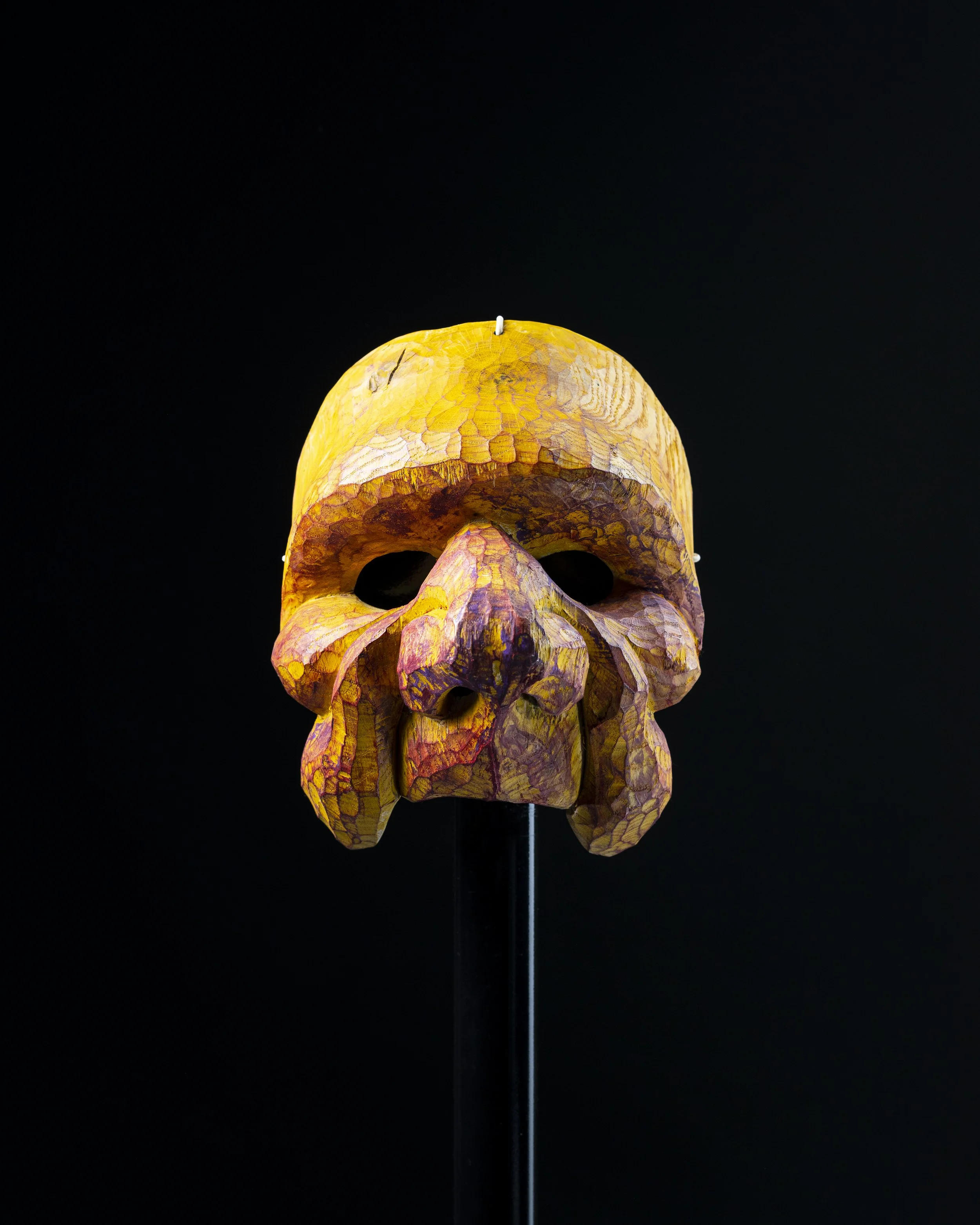 Masque en forme de crâne en bois peint en jaune et violet, exposé sur un support, fond noir.