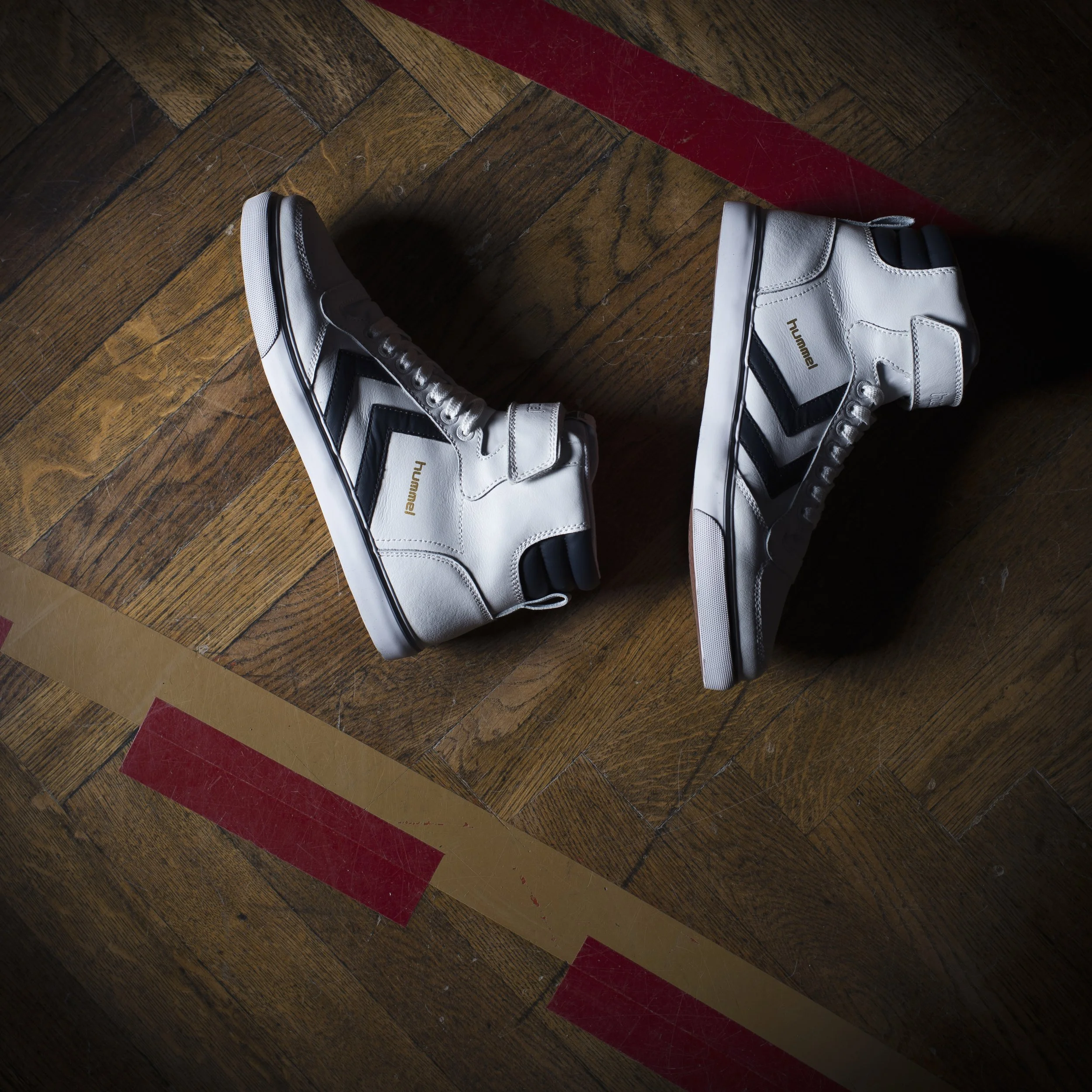 Des chaussures de sport blanches avec des accents noirs et dorés sur un sol en bois avec une ligne rouge.