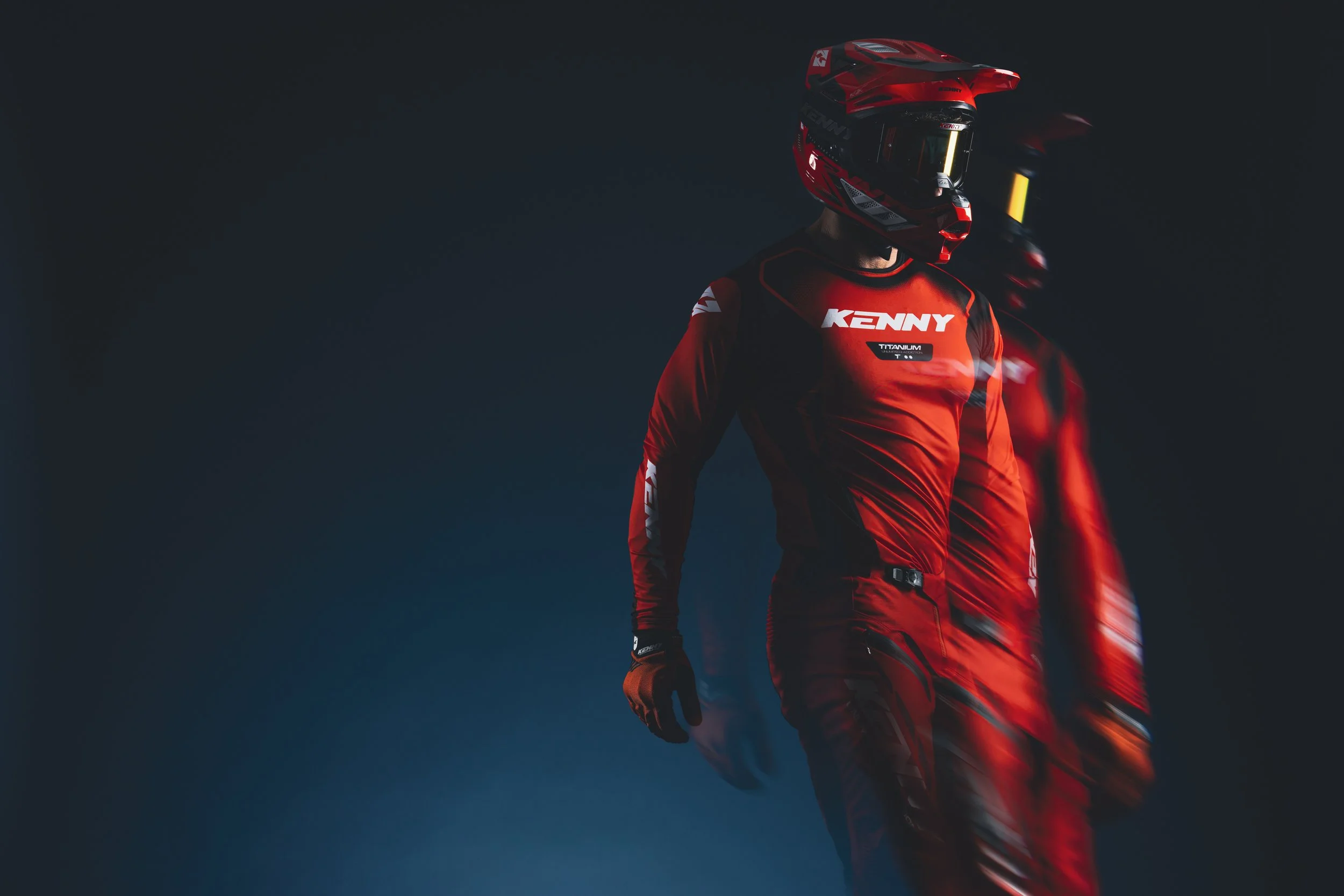 Deux motocross en tenue rouge et casque, debout dans un environnement sombre.