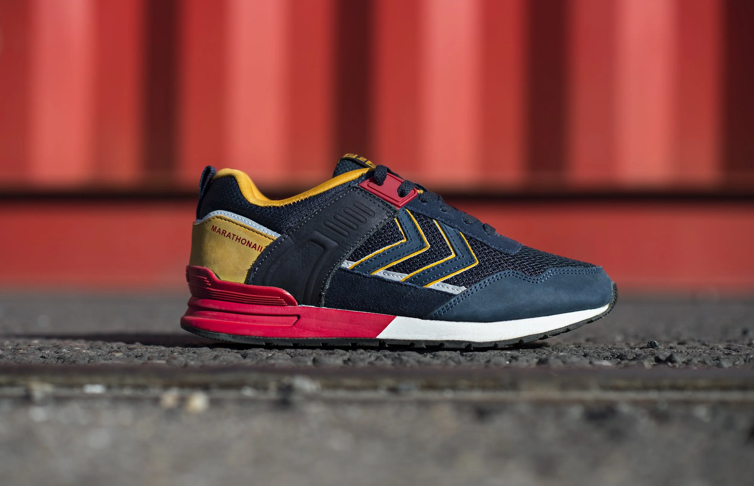 Chaussure de sport bleue, noire et rouge avec des détails jaunes et un talon marqué marathona, posée sur un sol asphalté, avec un fond rouge et noir.