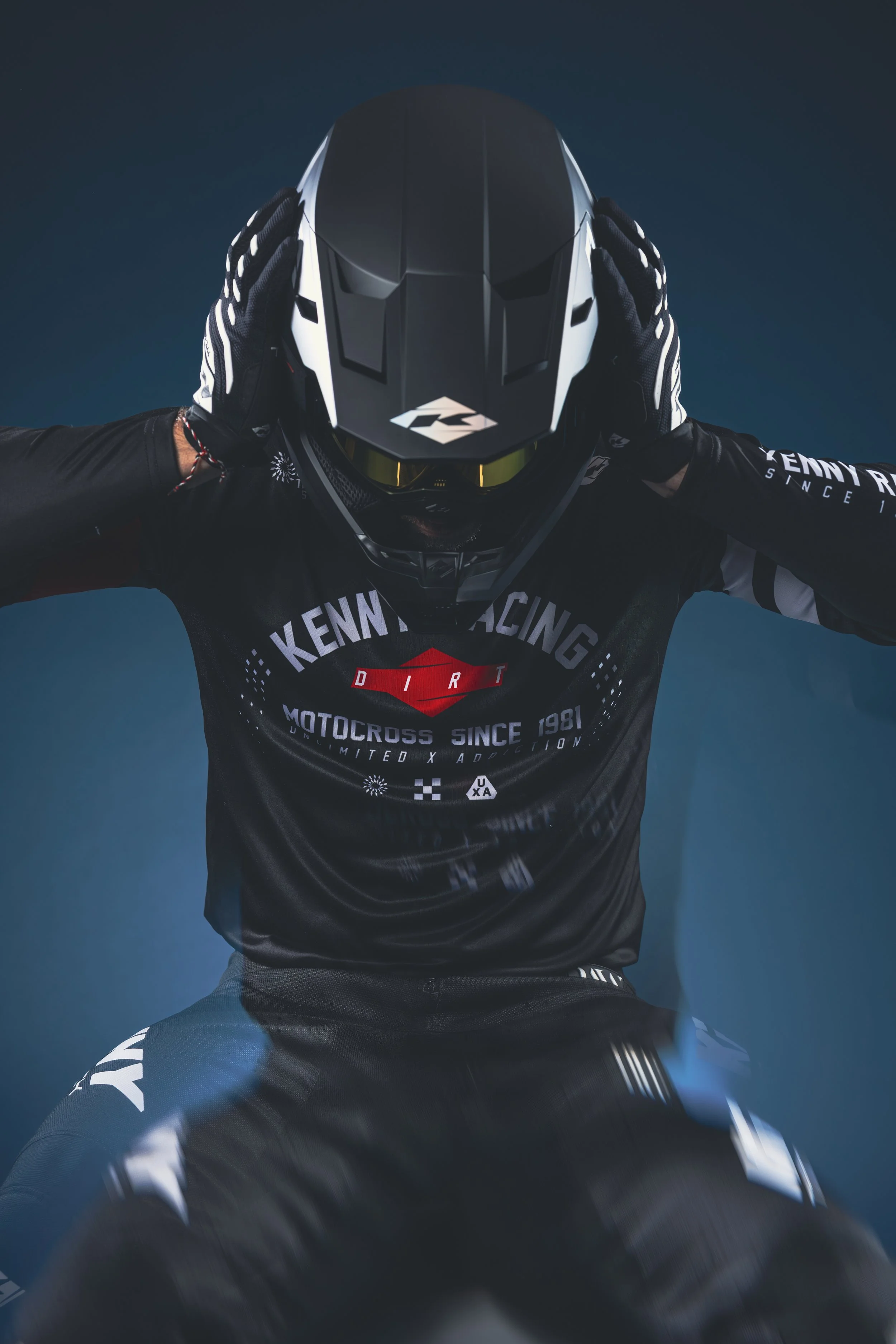 Un motocrossman en plein saut, portant un casque noir et blanc, une tenue noire avec des inscriptions blanches, sur un fond bleu.