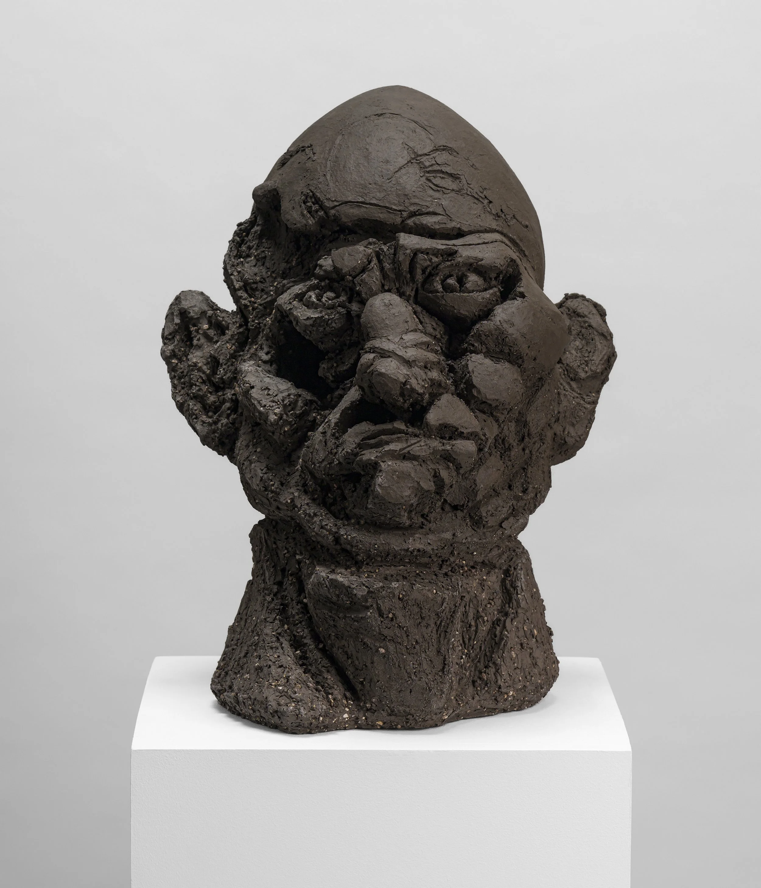 Sculpture en bronze représentant un personnage humain avec une tête disproportionnée, les mains sur le visage, exposée sur un socle blanc.