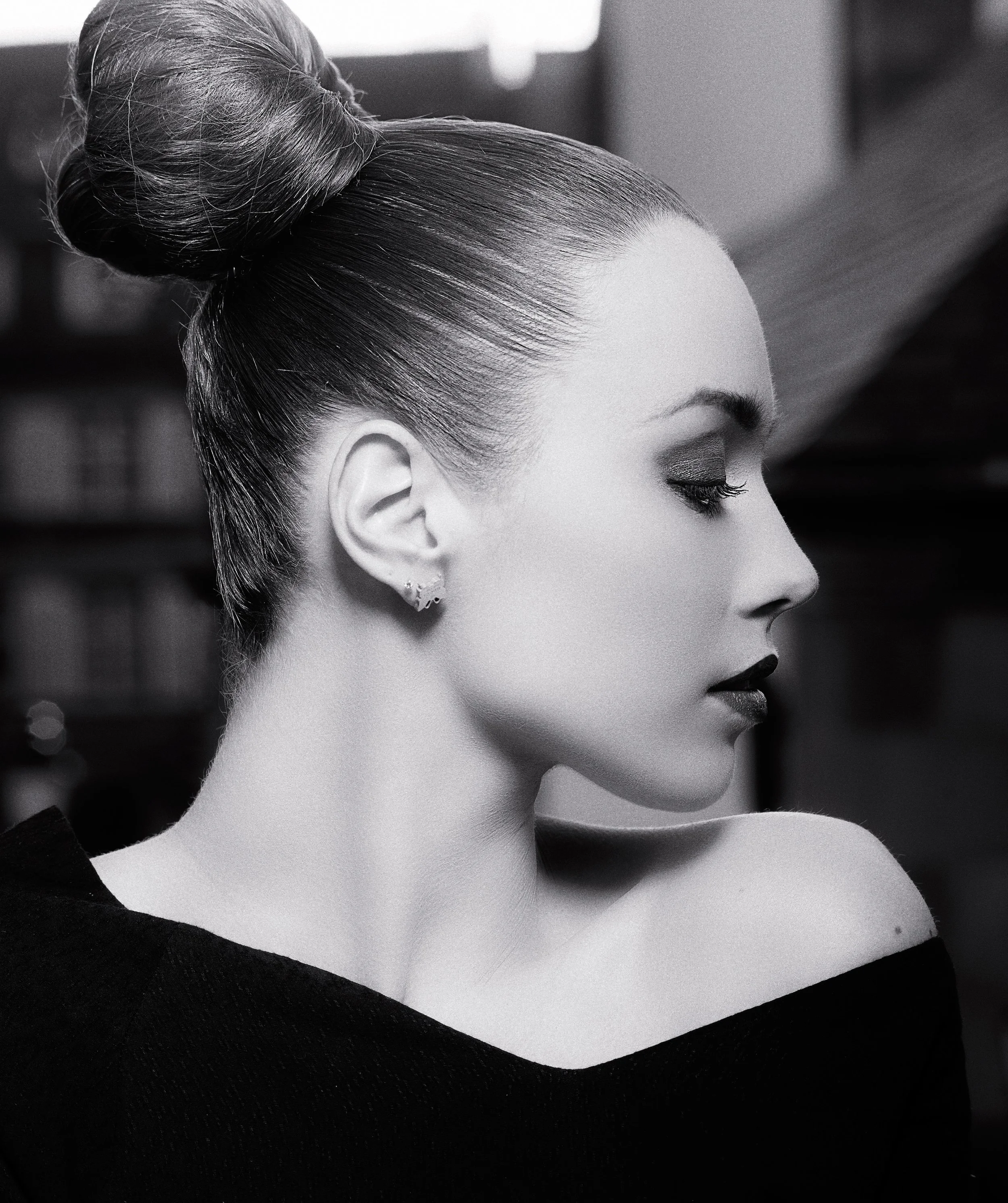 Une femme aux cheveux coiffés en chignon sophistiqué, vu de profil, portant des boucles d'oreilles, maquillée avec précision, vêtue d'une tenue sombre, dans un cadre élégant.