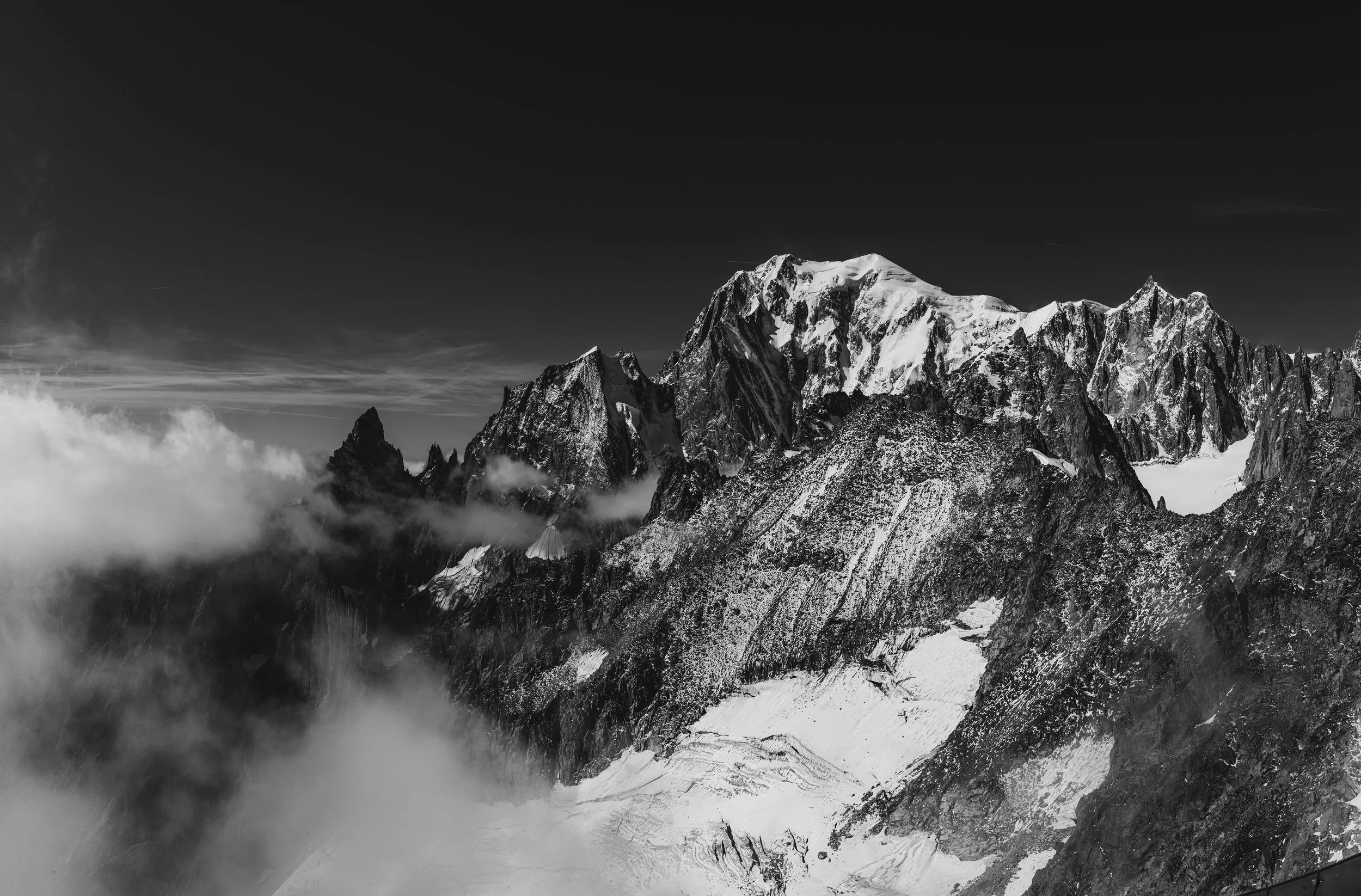 Montagne enneigée avec un sommet élevé, entourée de nuages et de falaises rocheuses, en noir et blanc