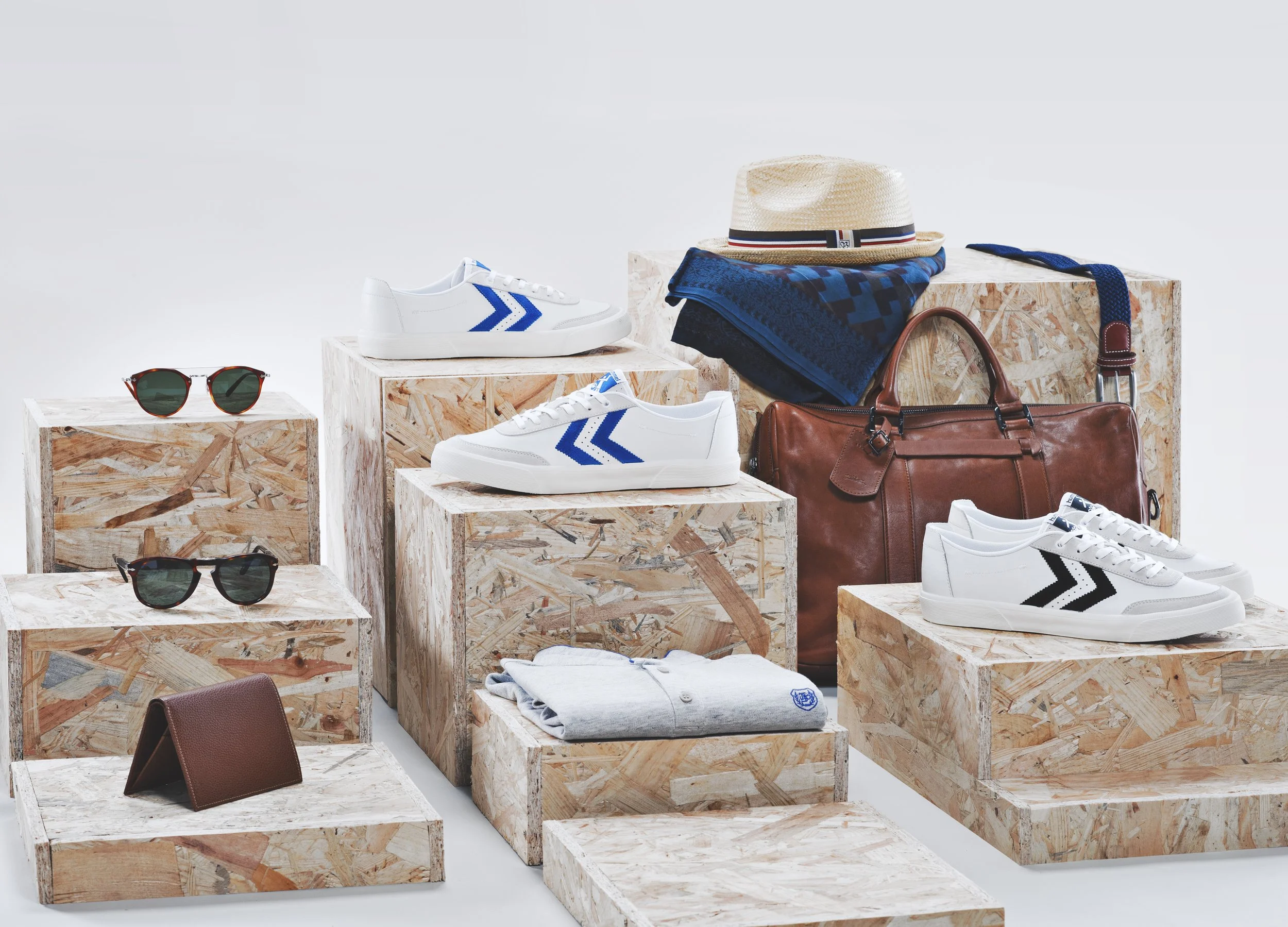 Collection d'accessoires de mode comprenant des lunettes de soleil, des baskets blanches avec des détails bleus, un sac en cuir marron, un chapeau en paille, des foulards, une ceinture, et des vêtements en coton, tous disposés sur des placages en boi