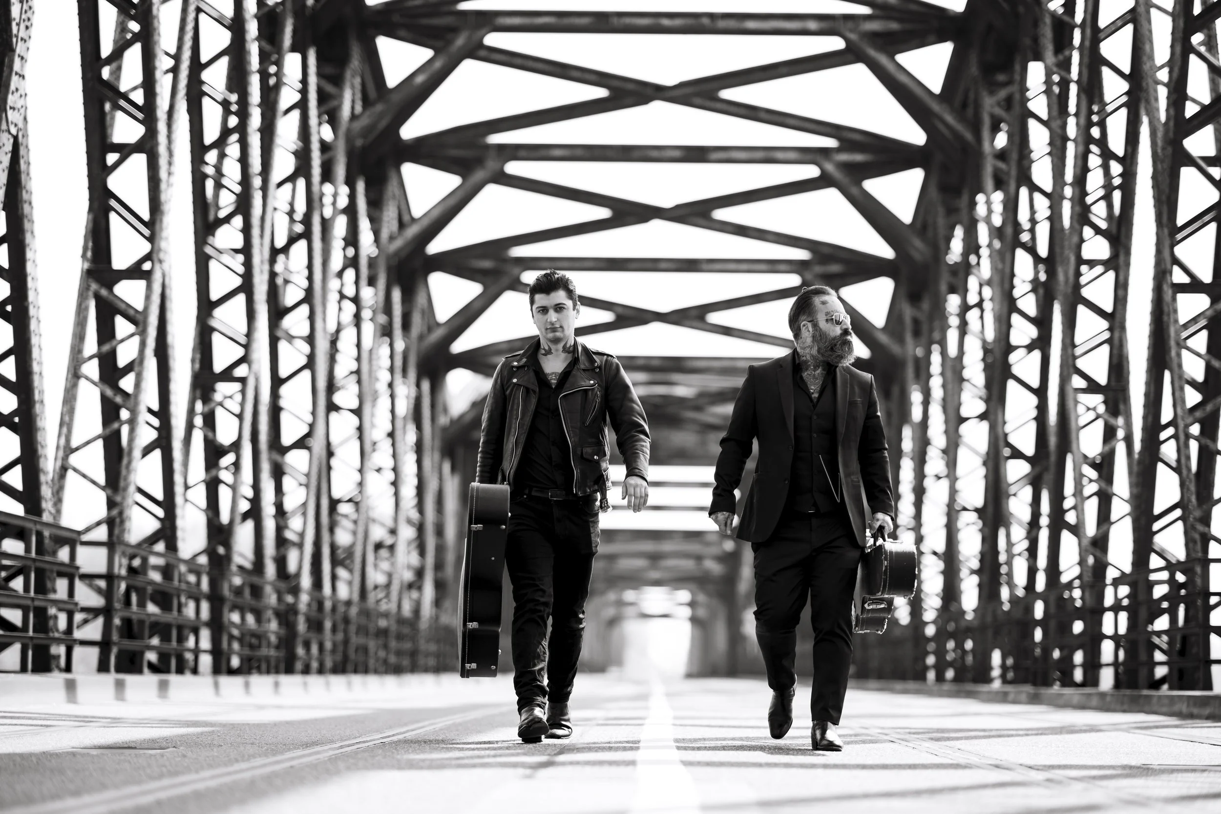 Deux hommes marchent sur un pont métallique en noir et blanc, chacun avec une valise. L'un porte une veste en cuir, l'autre un costume avec une barbe et des lunettes. Le pont a une structure en acier complexe.