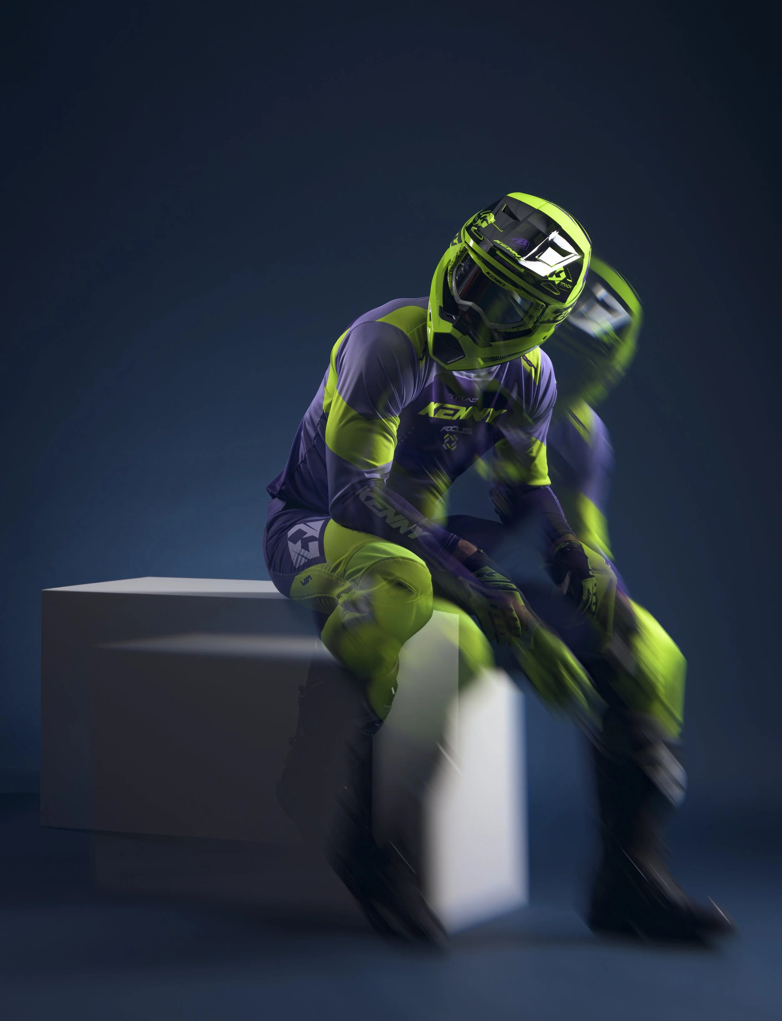 Un motocycliste en tenue de protection et casque de couleur jaune et violet, en position de moto-cross, sur un fond sombre.