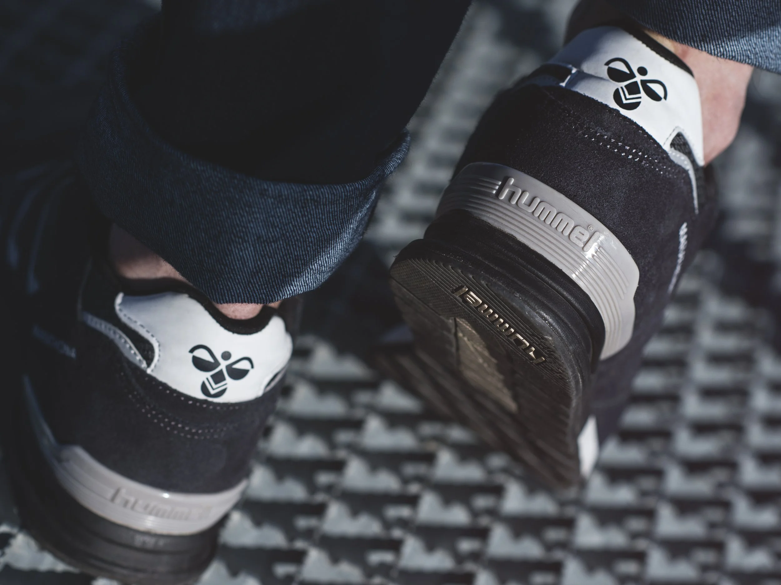 Chaussures de marche noires et blanches avec des logos, portées par une personne dont une jambe est partiellement visible, marchant sur une surface métallique à relief - marque HUMMEL