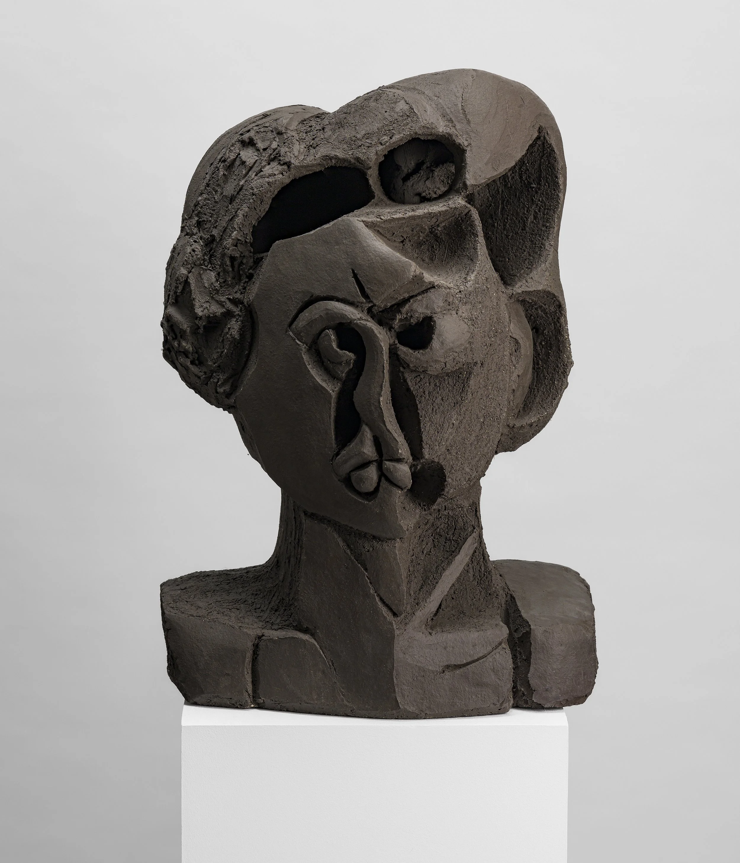 Sculpture abstraite en pierre gris foncé, représentant un visage stylisé avec des détails simplifiés, sur une base blanche.