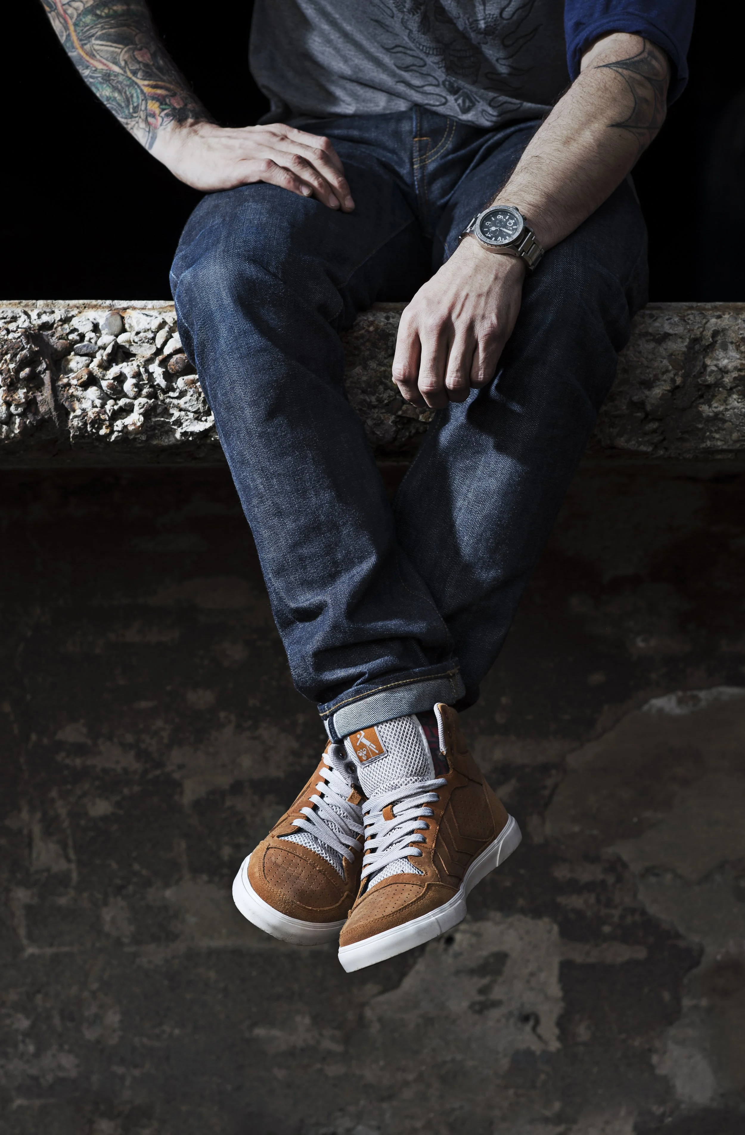 Seated person wearing a blue hoodie, jeans, and brown sneakers, with visible tattoos on arms and wristwatch, on a stone surface.