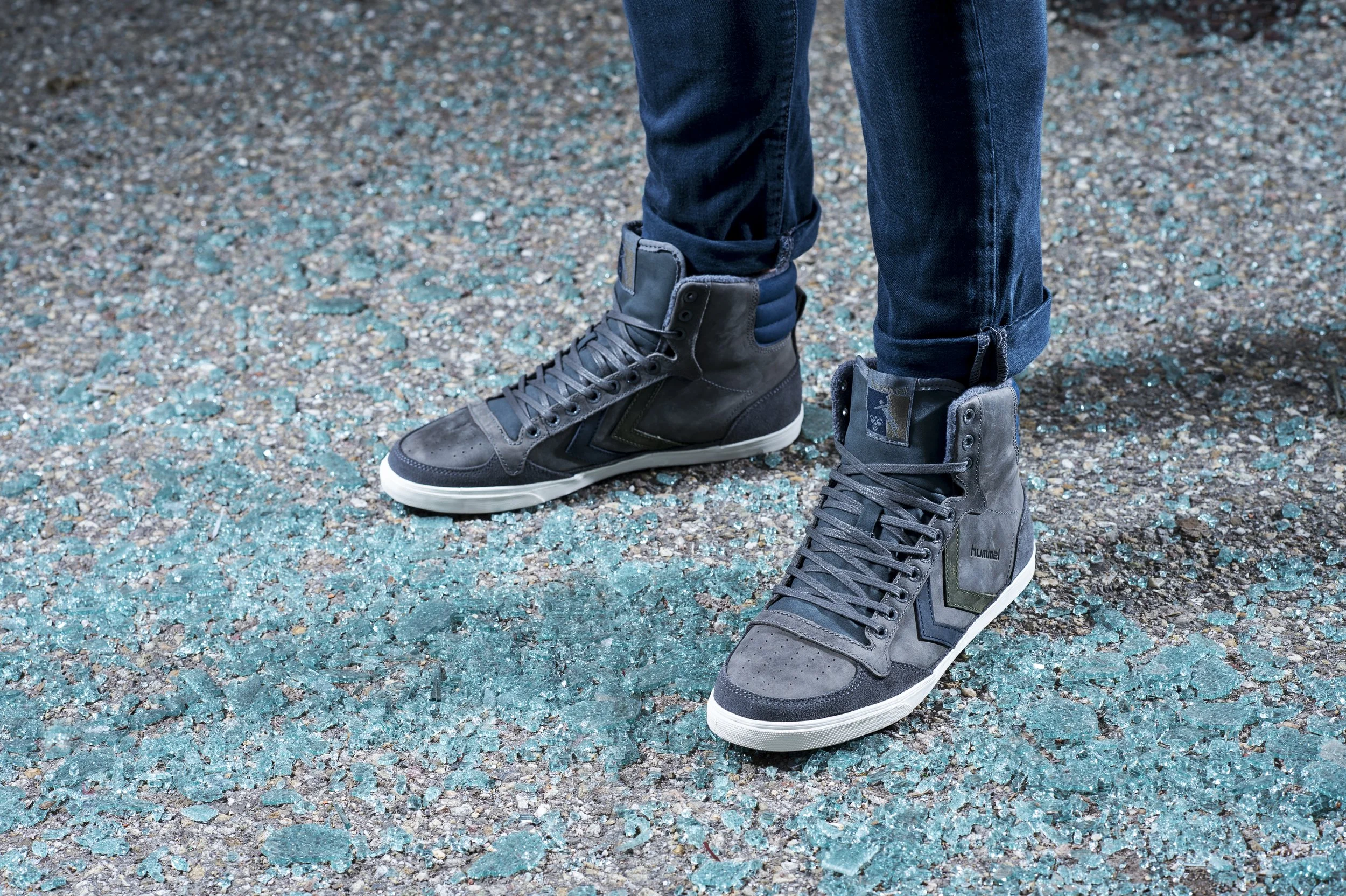 Chaussures de sport grises avec des détails noirs et verts, portées par une personne en jean, debout sur un sol de graviers avec des éclats de verre bleu. La personne porte des pantalons en jean roulés à la cheville.
