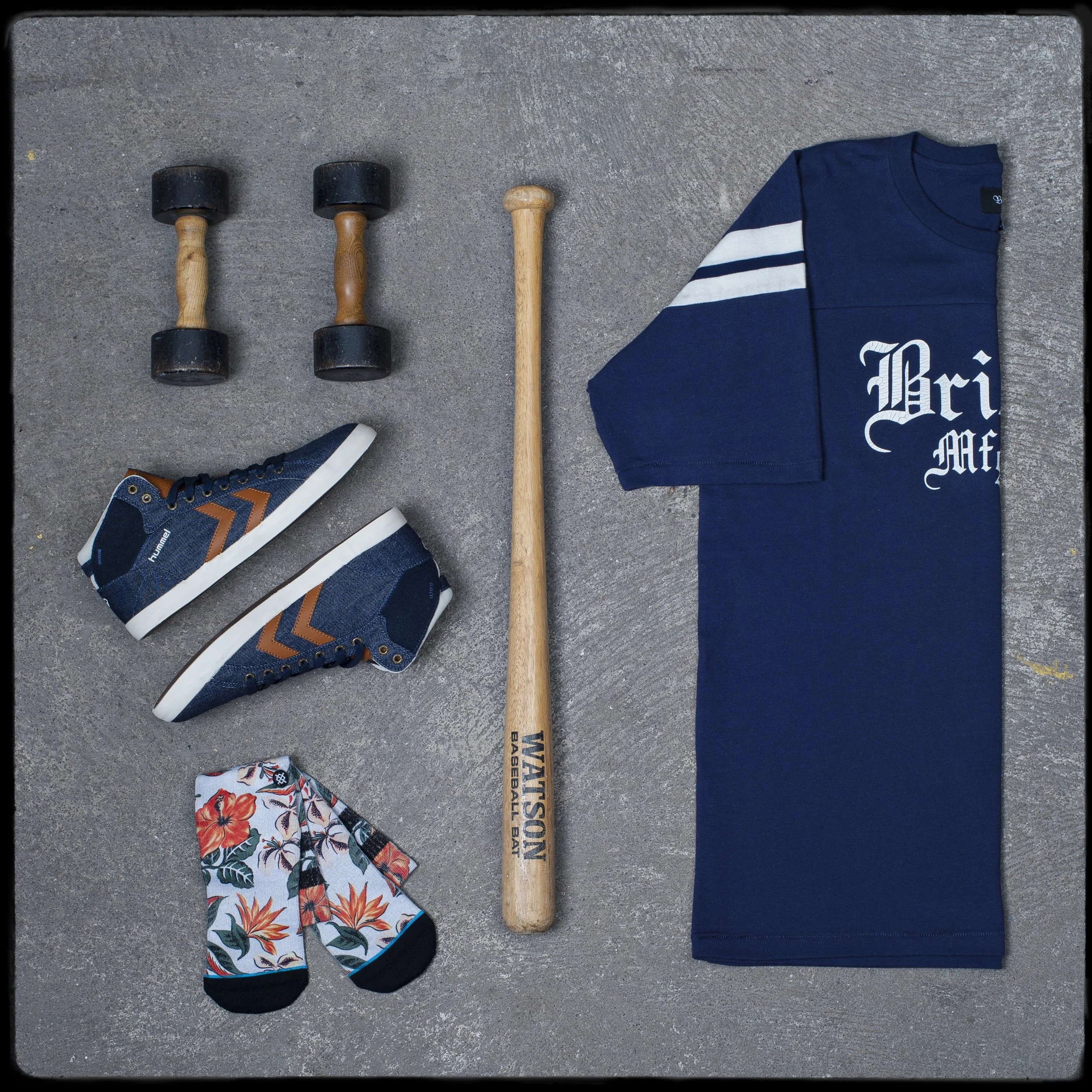 Une paire de baskets de sport noires avec des accents orange, une paire de chaussettes à motif floral, une cotte de mailles bleue foncée, un t-shirt bleu avec écriture blanche, un marteau en bois et une pièce de tissu colorée sont disposés sur une su