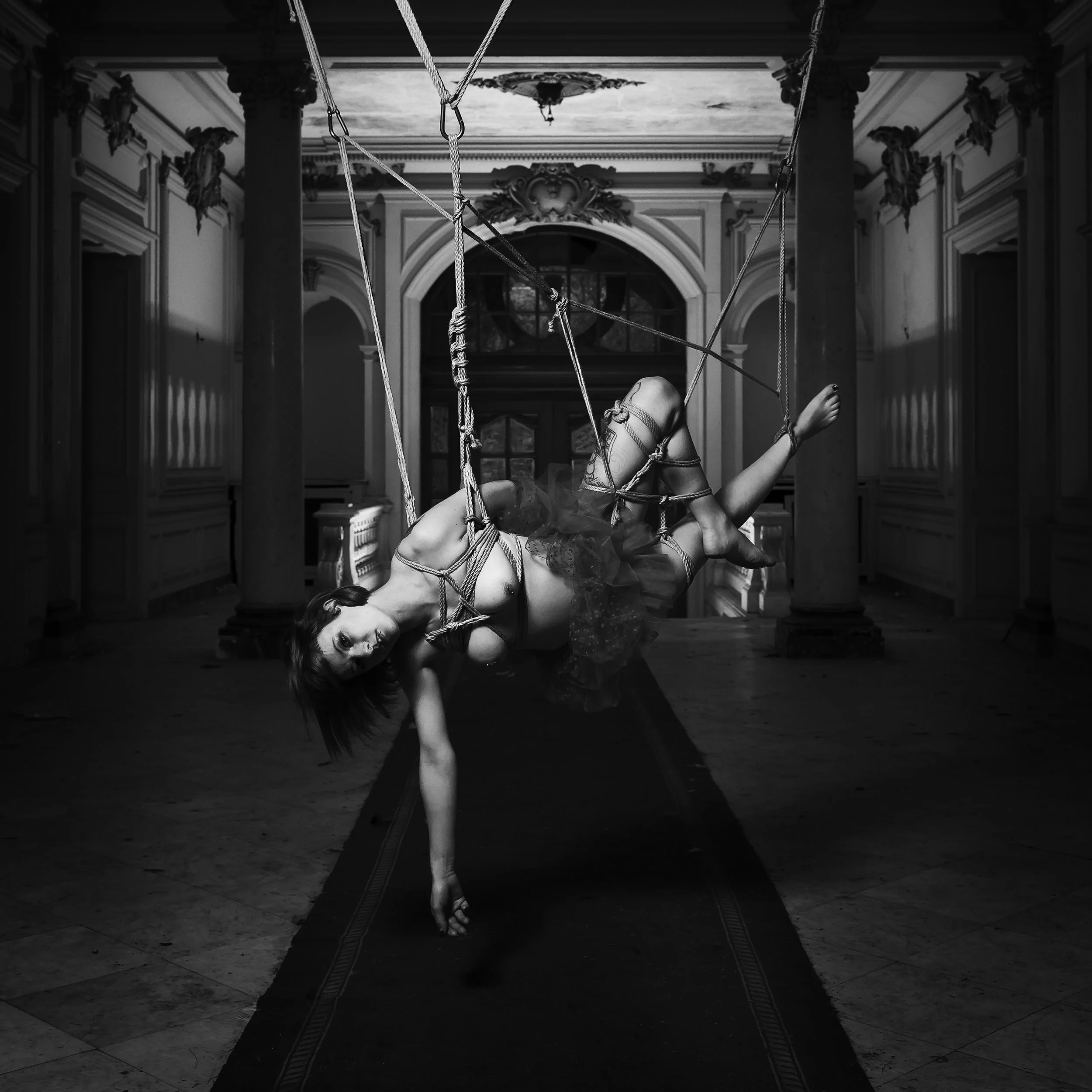Une sculpture de femme nue allongée dans une pièce majestueuse, entourée de cordages attachés à ses poignets, chevilles et tête, dans un style artistique sombre et mystérieux.
