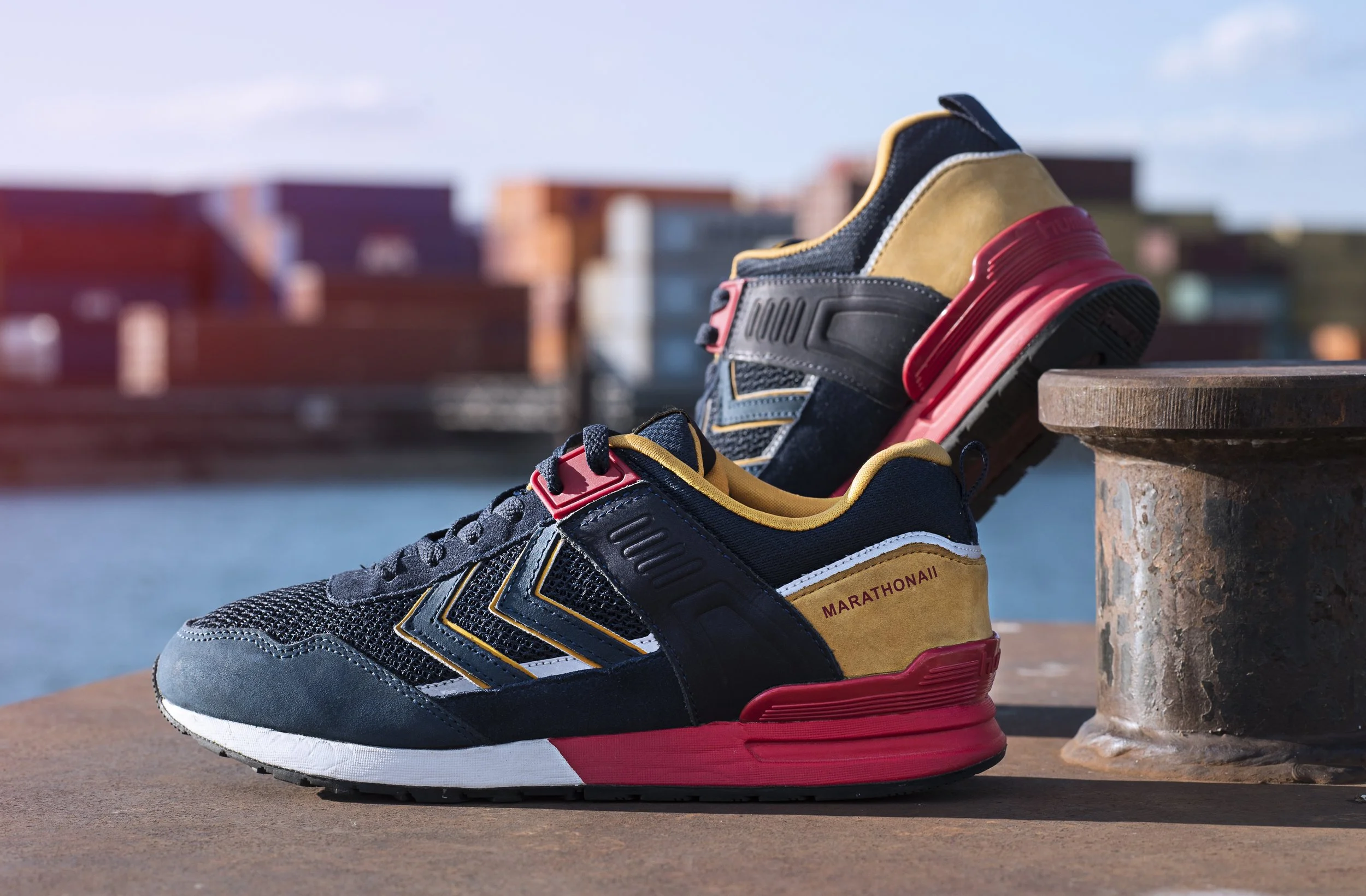 Une paire de chaussures de course colorées posée sur une surface en béton à côté d'une colonne en pierre, avec un arrière-plan flou d'immeubles modernes et un ciel partiellement nuageux.