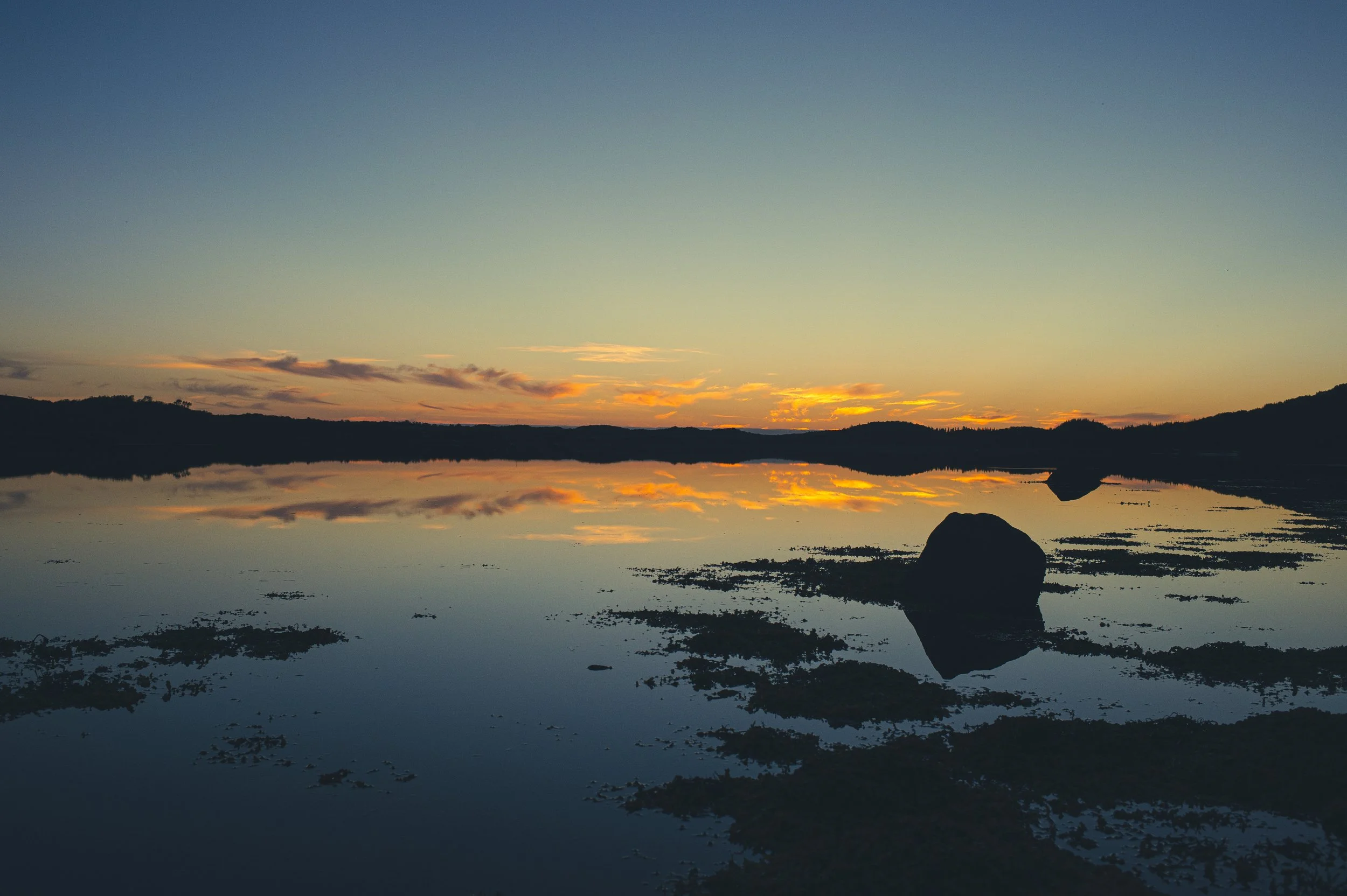 Coucher de soleil sur un lac calme avec des montagnes en arrière-plan, reflet des nuages orange et jaune sur l'eau.