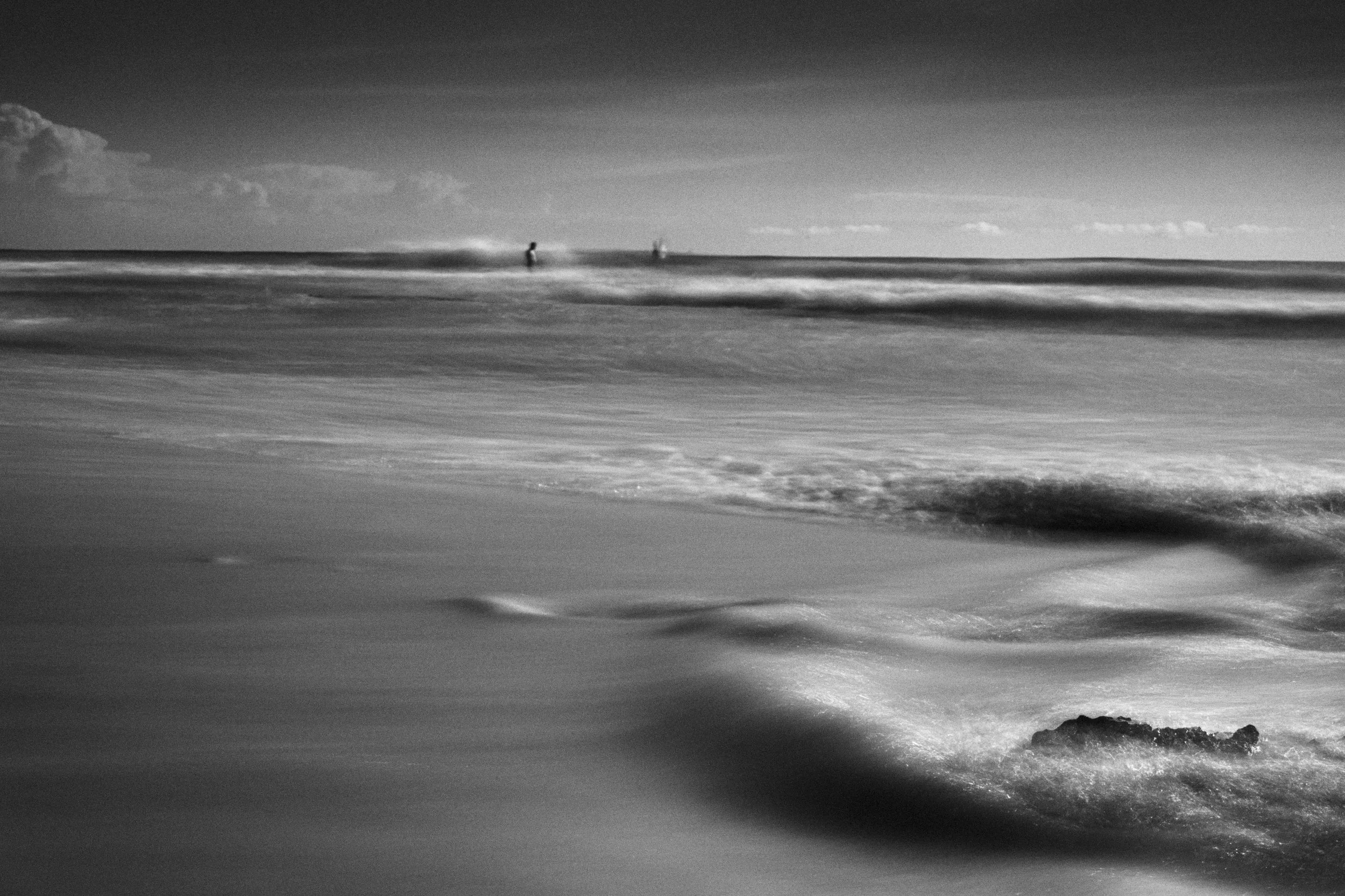 Plage déserte en noir et blanc avec des vagues et deux personnes au loin sur l'horizon.