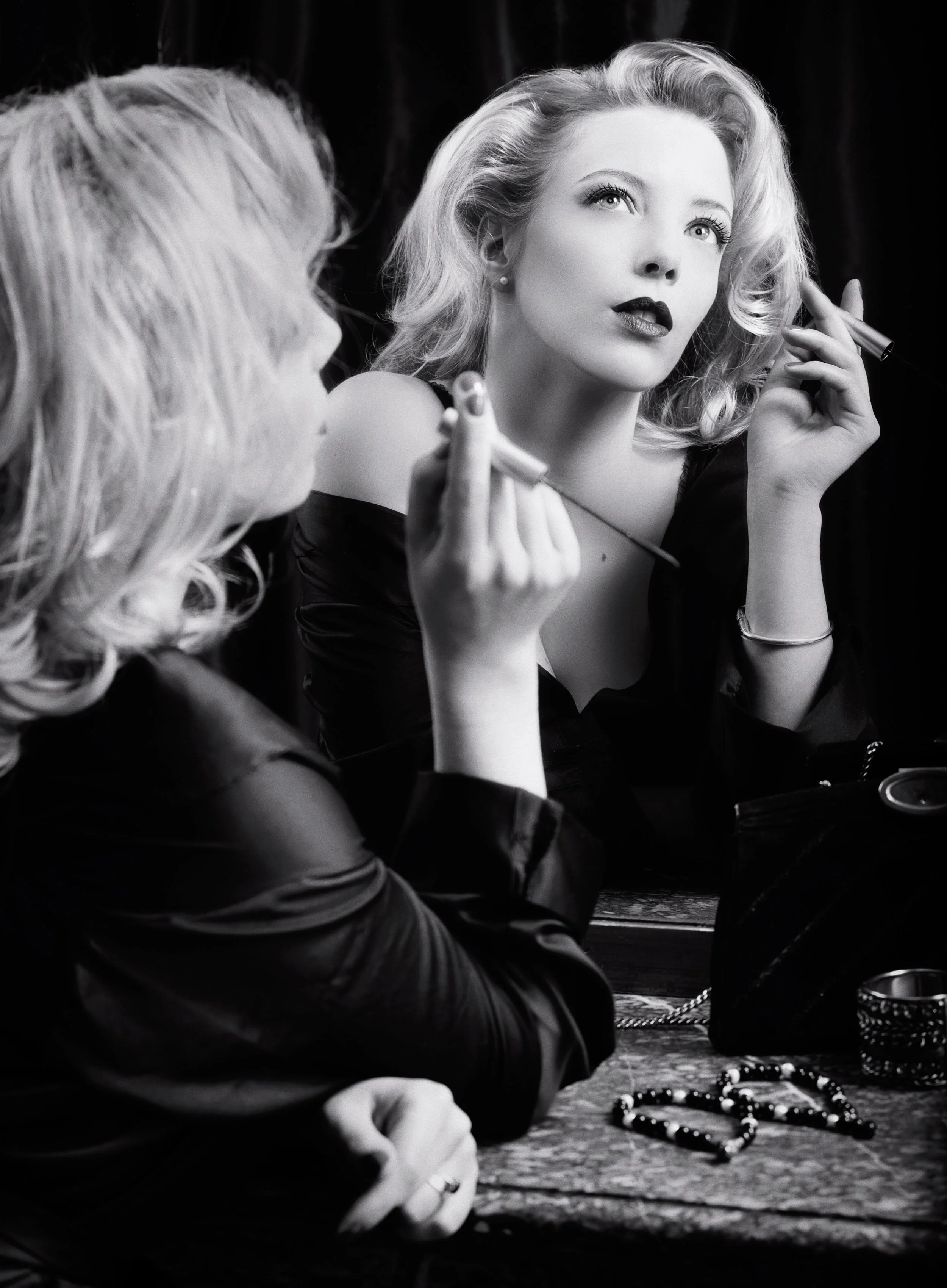 Une femme blonde regardant pensivement, avec du maquillage foncé, en train de se faire coiffer ou maquiller par une autre personne, en noir et blanc.