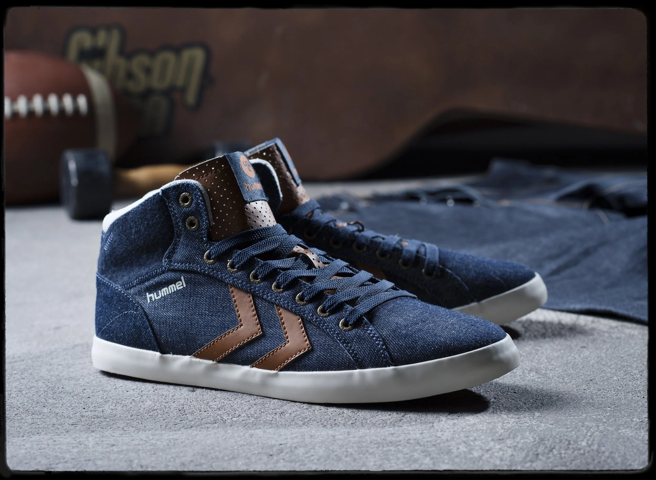 Une paire de baskets à lacets bleus avec des bandes marron, posées sur un sol en béton avec un ballon de football flou en arrière-plan.