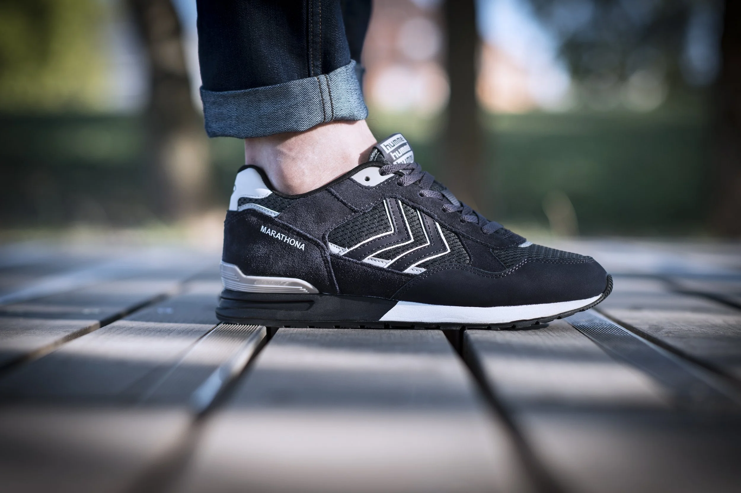 Une chaussure de course noire avec des détails blancs et la marque "Hummel" sur la languette, portée par une personne dont le pantalon est roulé au bas, marchant sur un pont en bois en plein air.