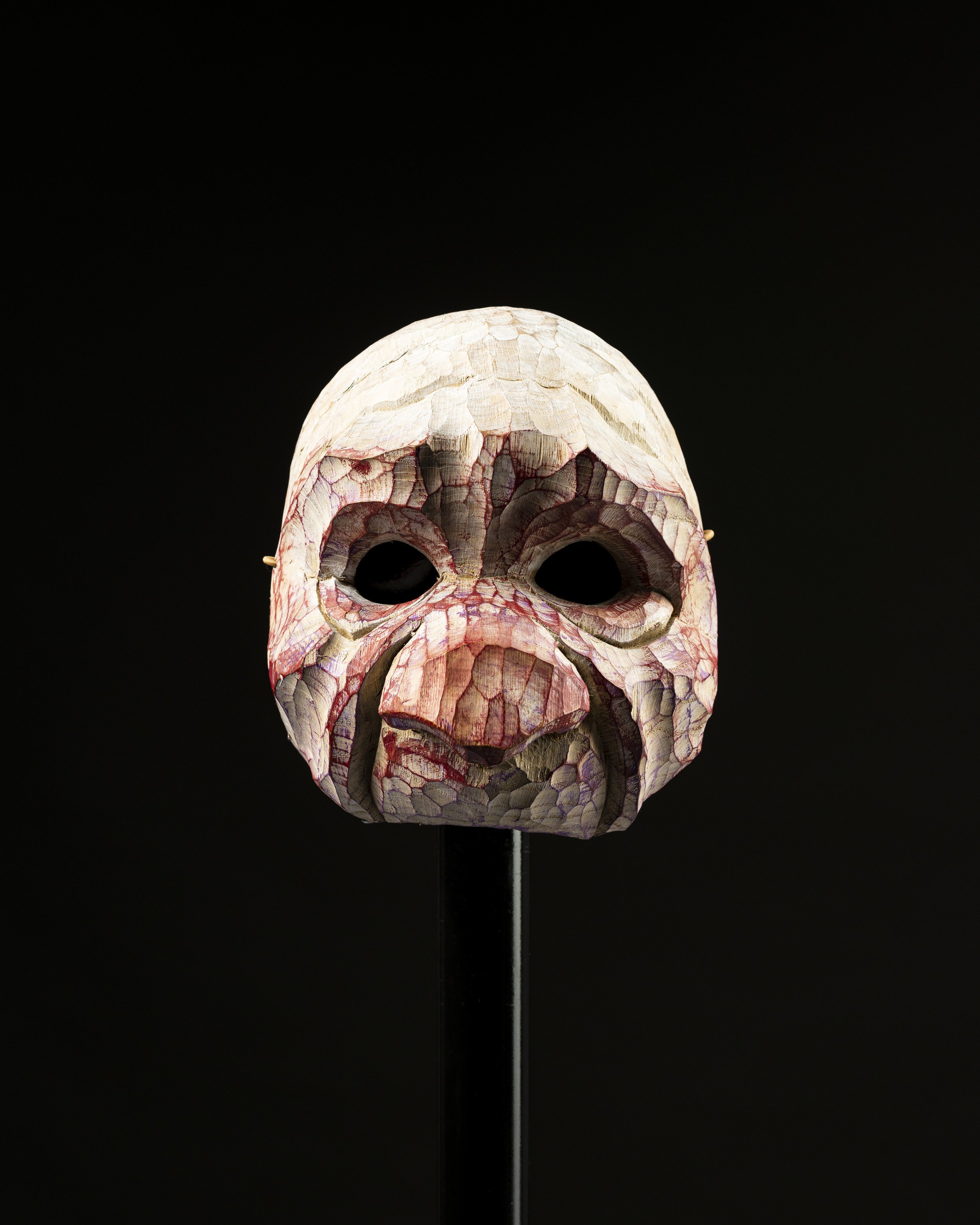 Masque en papier mâché représentant un visage humain avec des détails en relief et des couleurs naturelles, exposé contre un fond noir.