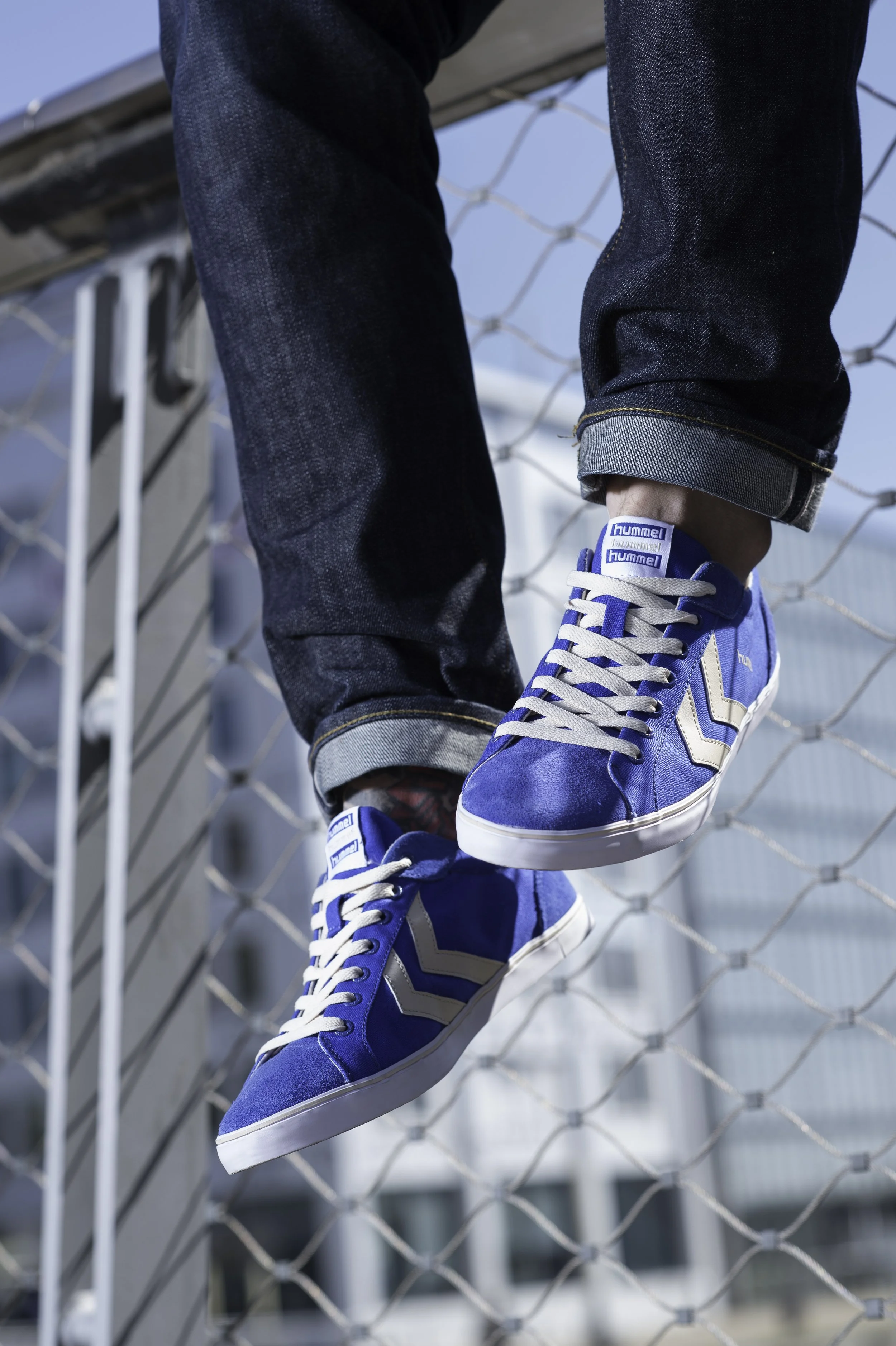 Une paire de chaussures sneakers bleues avec des lacets blancs, portée par une personne dont le pantalon est retroussé, en train de marcher sur une clôture en fil de fer.
