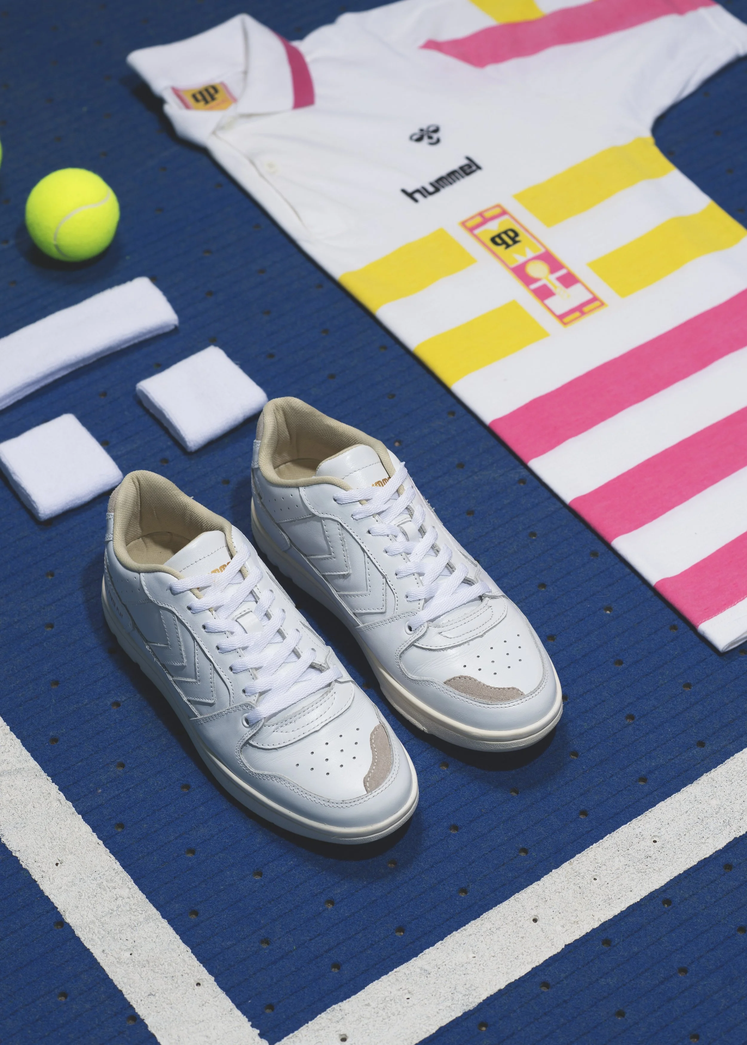 Vêtements et équipement de tennis sur un court en bleu, comprenant une paire de baskets blanches, une balle de tennis jaune, une serviette blanche, un tee-shirt blanc avec un logo et des écritures, et trois bandes de tissu blanc - marque HUMMEL