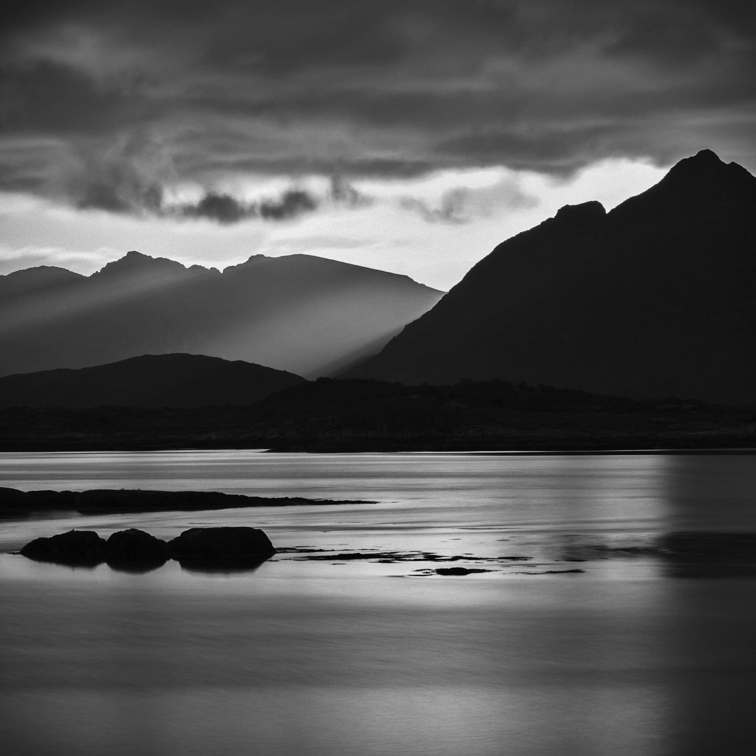 Paysage de montagnes en noir et blanc avec un ciel nuageux et un lac calme au premier plan.