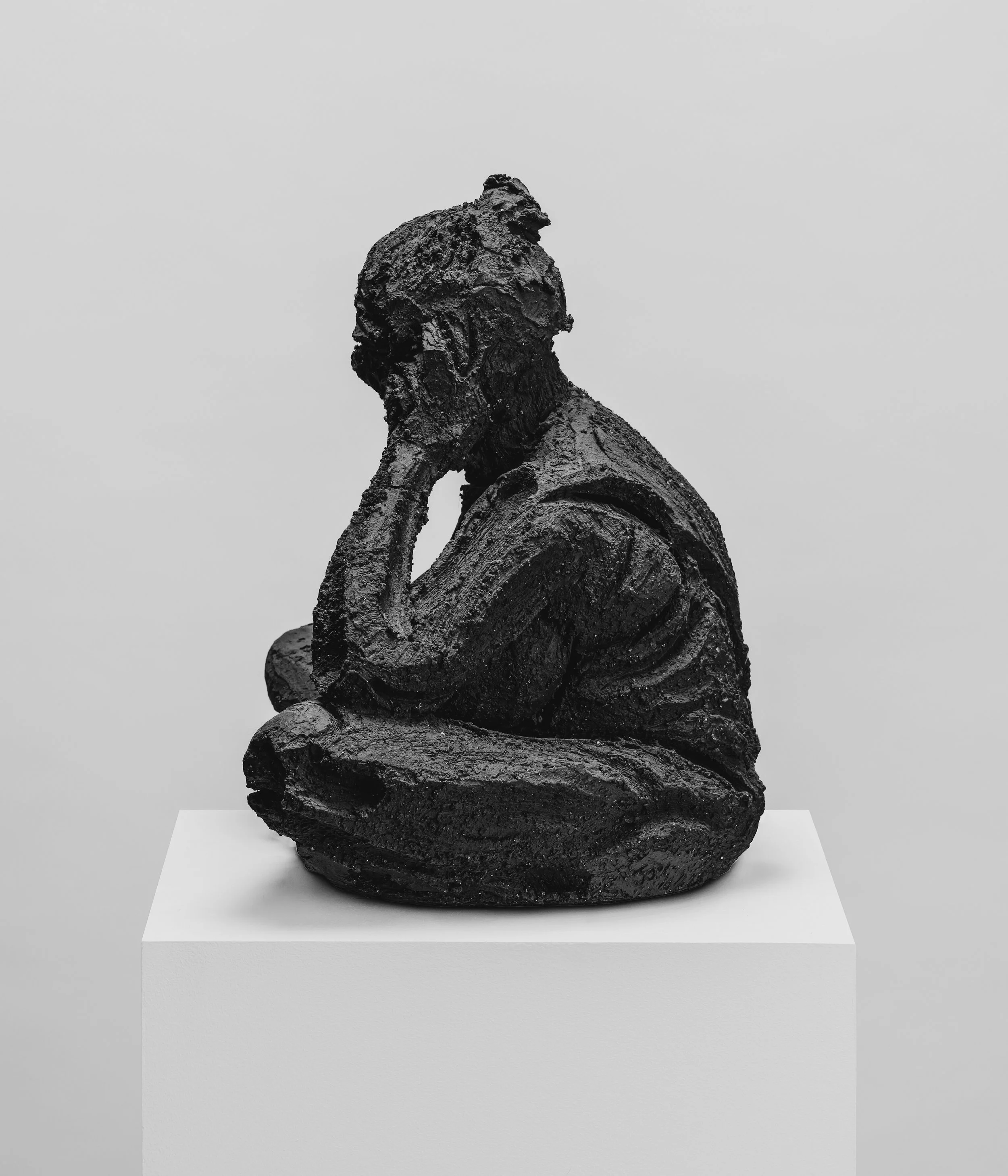 Sculpture en bronze représentant une figure humaine assise, les mains sur le visage, exposée sur une plateforme blanche.