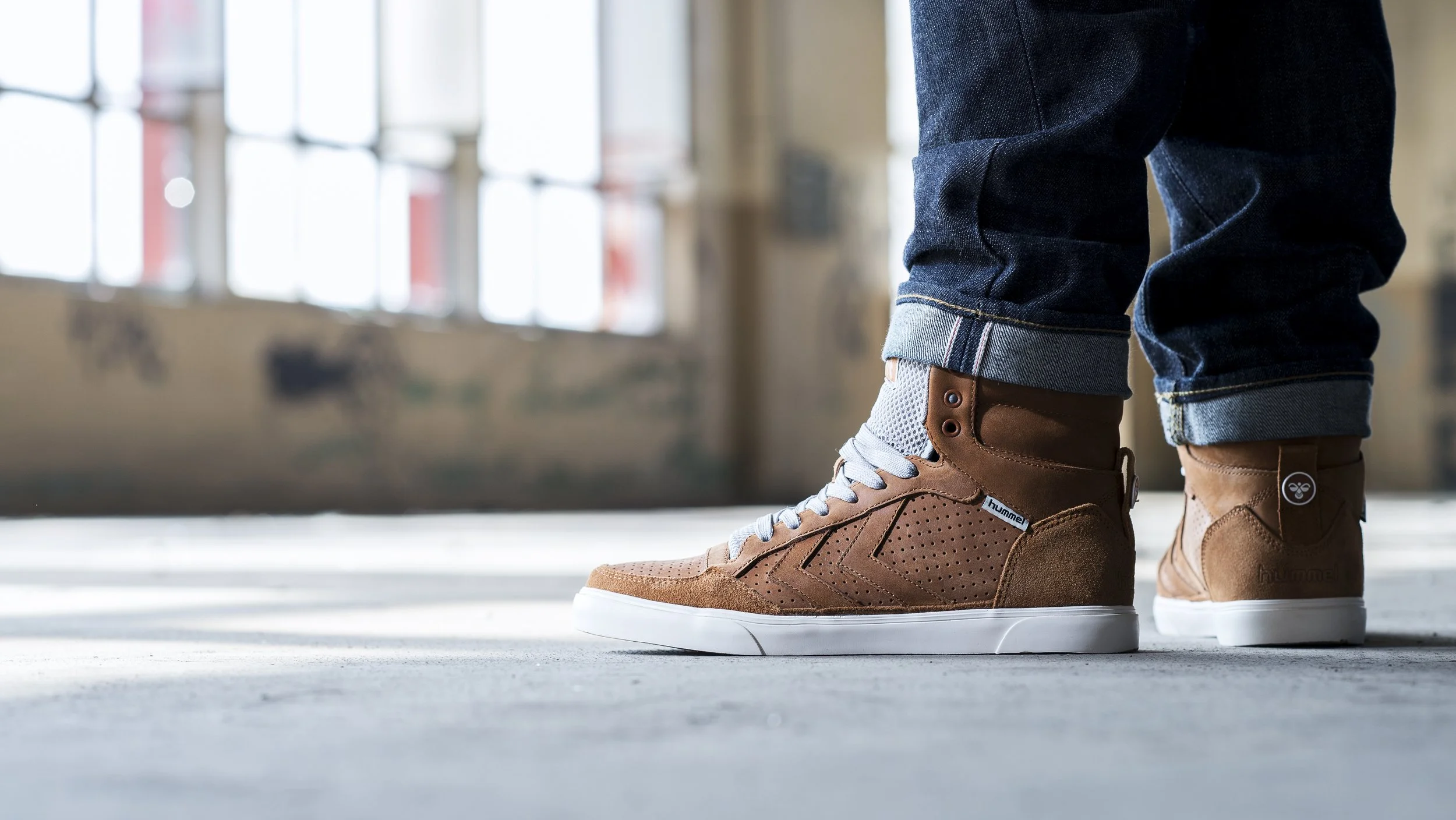 Personne portant des baskets marron de marque Hummel, avec des jeans roulés en finition et un fond d'espace industriel avec fenêtres