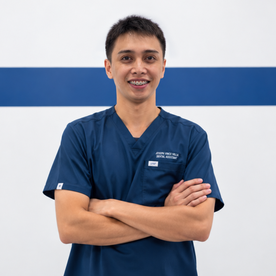 Young male dental assistant wearing navy scrubs, standing with arms crossed and smiling in front of a white and blue wall.