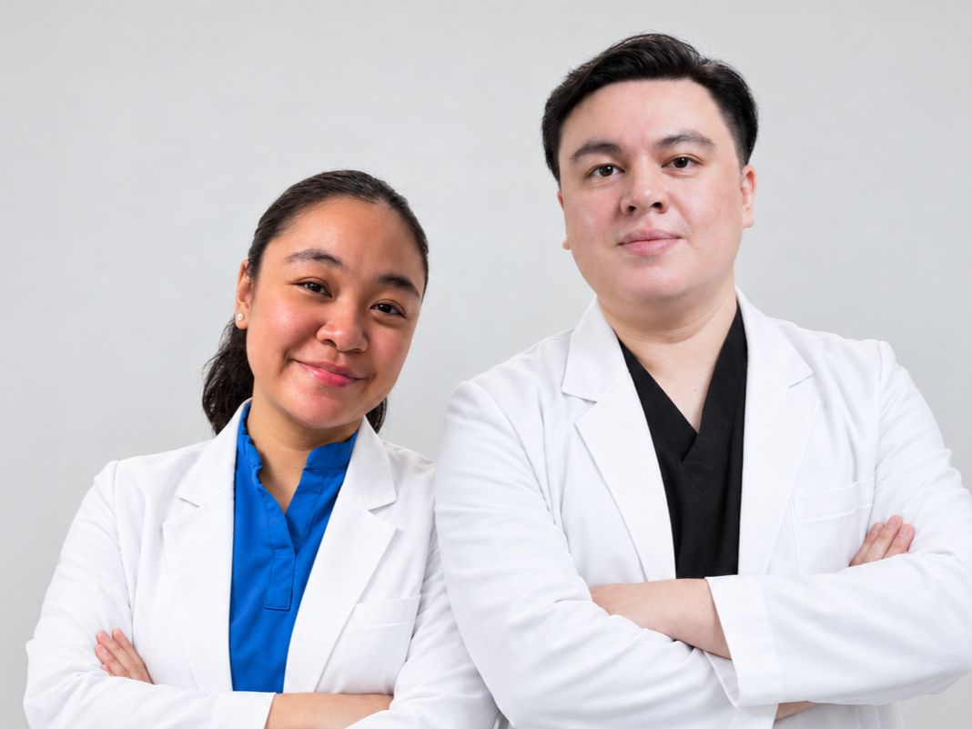 Two healthcare professionals, a woman and a man, standing with arms crossed in white lab coats against a plain light gray background.