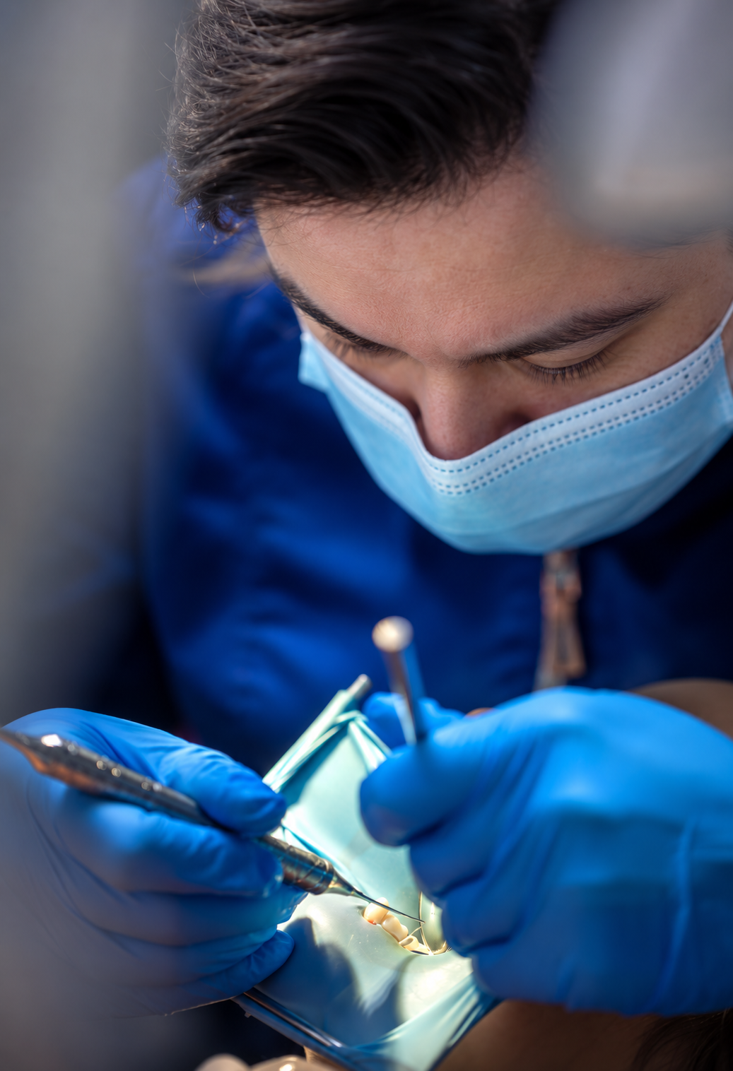 A person wearing a face mask and blue gloves is performing dental surgery, using dental tools inside a patient's mouth.