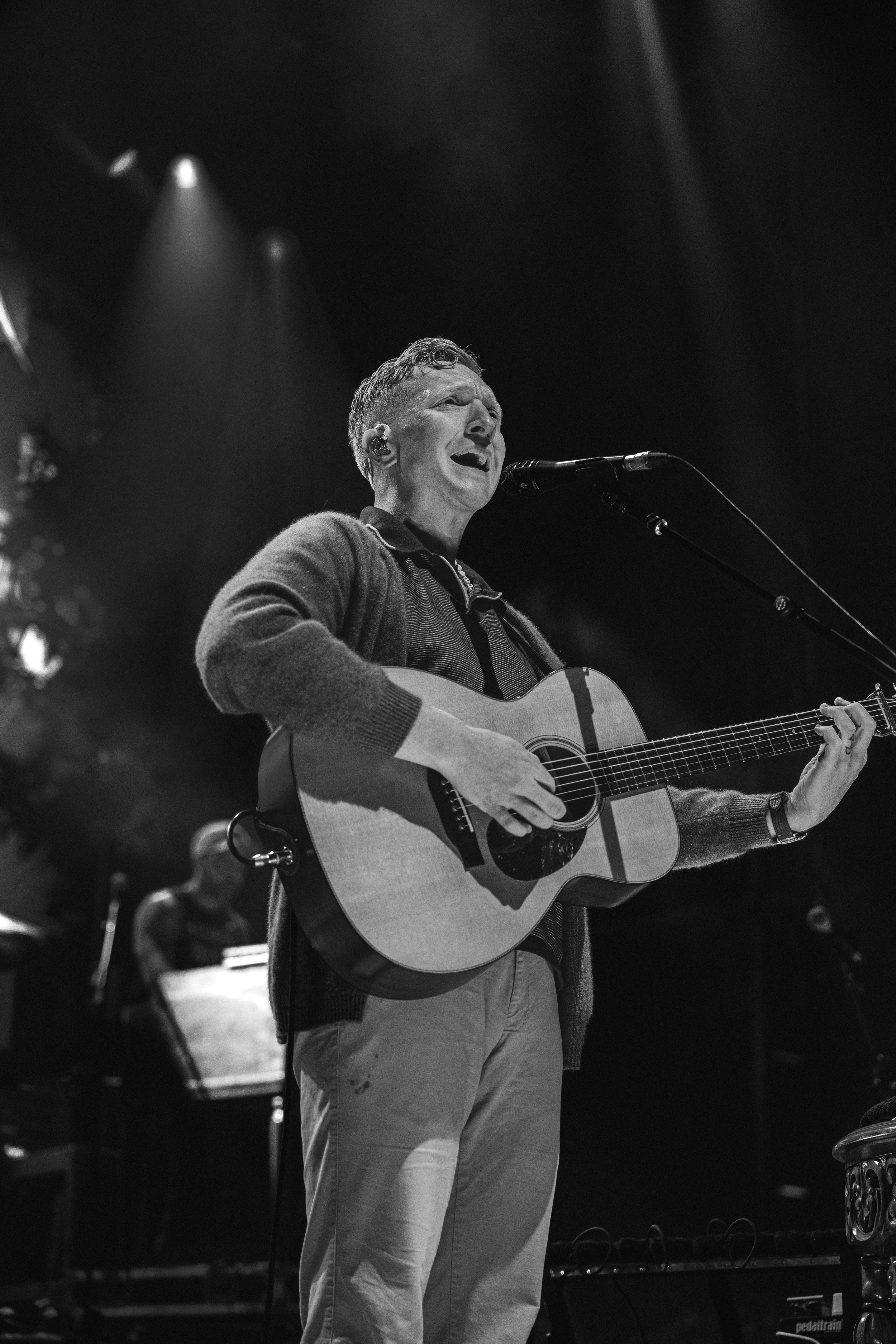 A man with short curly hair singing and playing an acoustic guitar on stage, illuminated by stage lights, with a microphone in front of him.