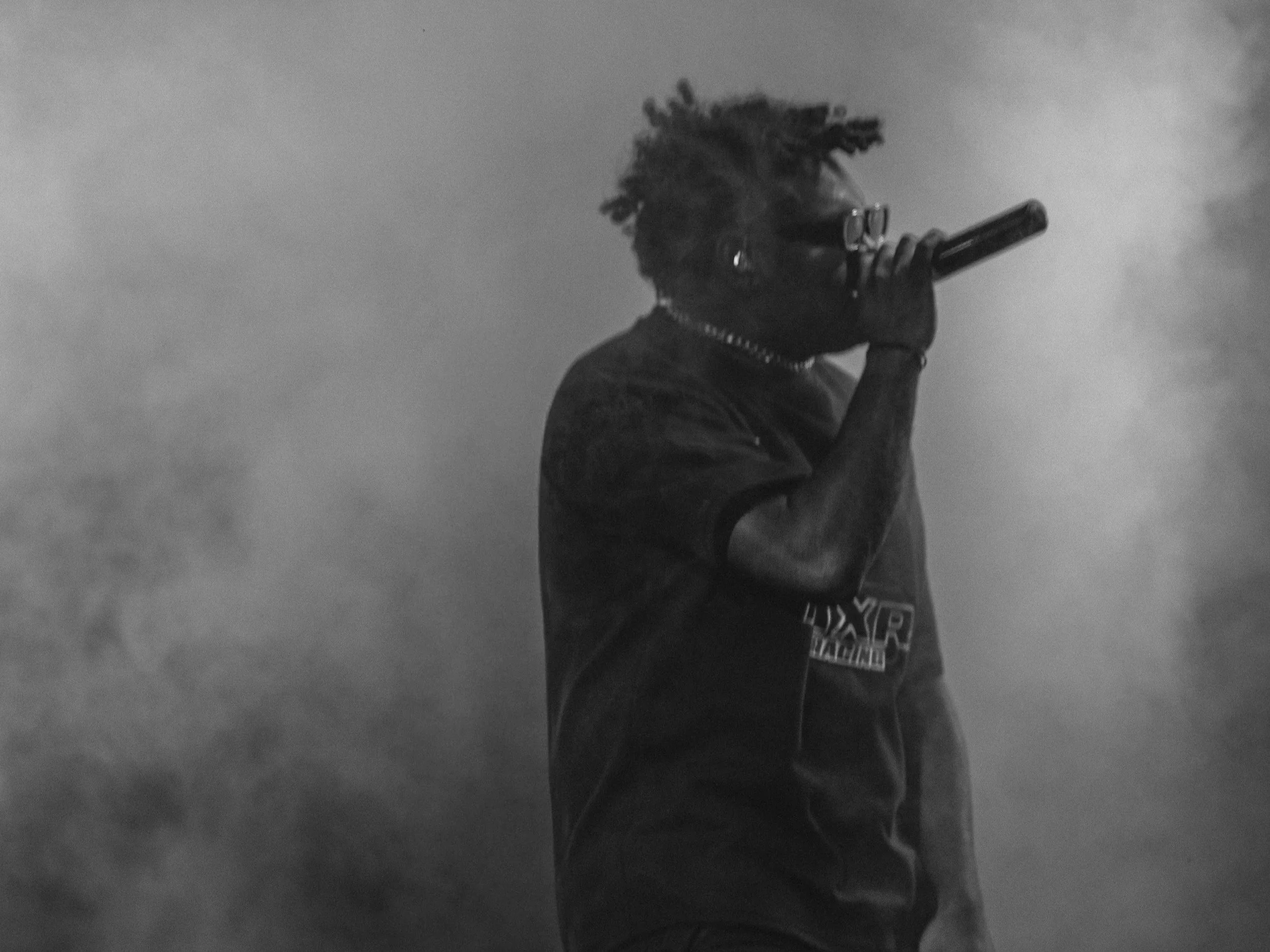 A person with dreadlocks and glasses holding a gun to their head, standing against a cloudy sky.