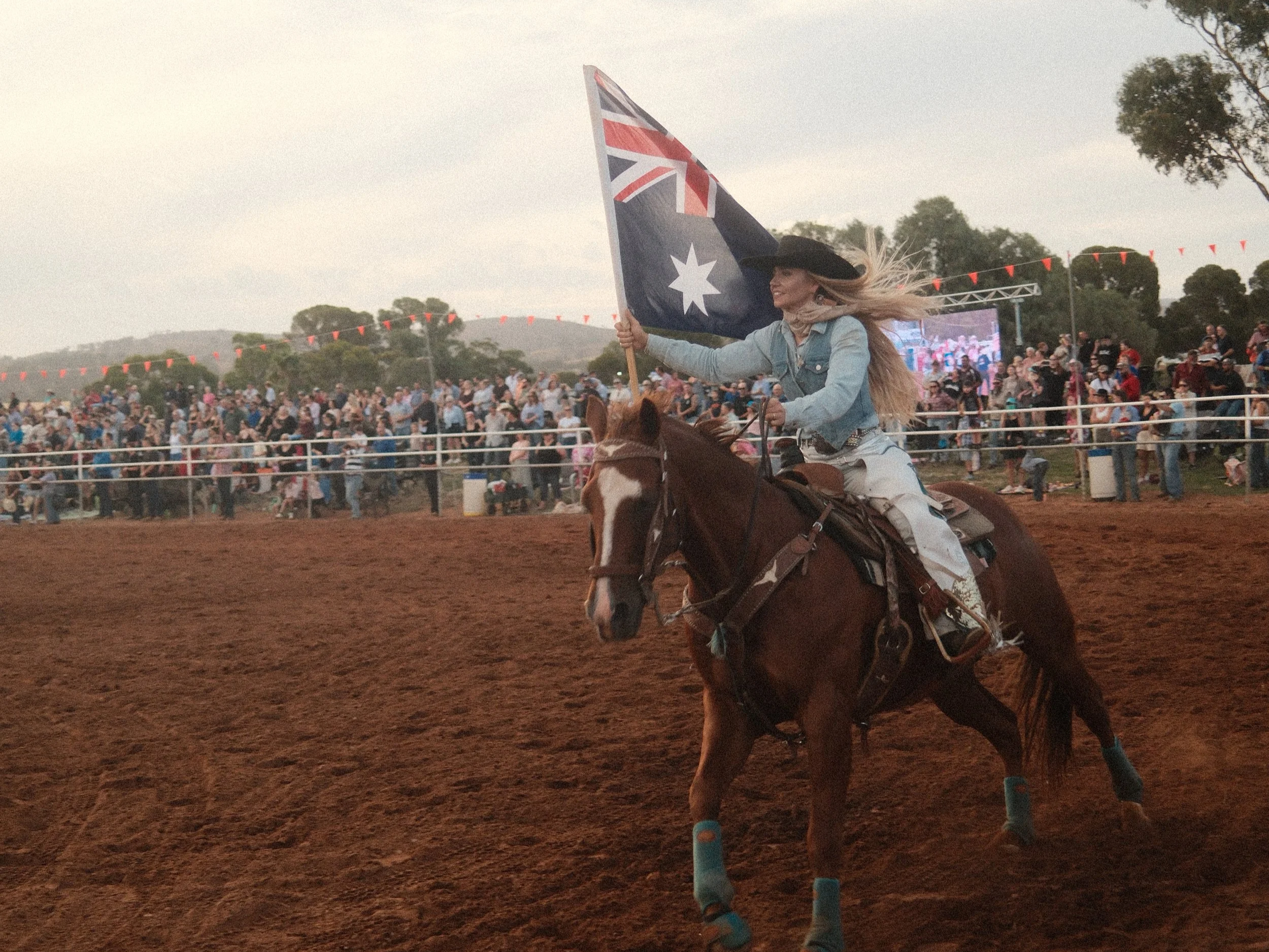 A woman riding a horse at an outdoor event, holding a large Australian flag, with a crowd and a big screen in the background.