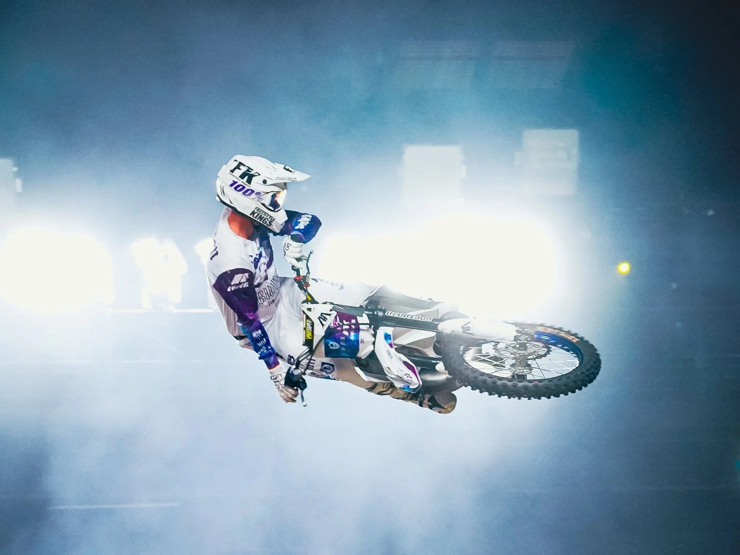 A person in motocross gear riding a dirt bike in a foggy or smoky environment with bright lights in the background.