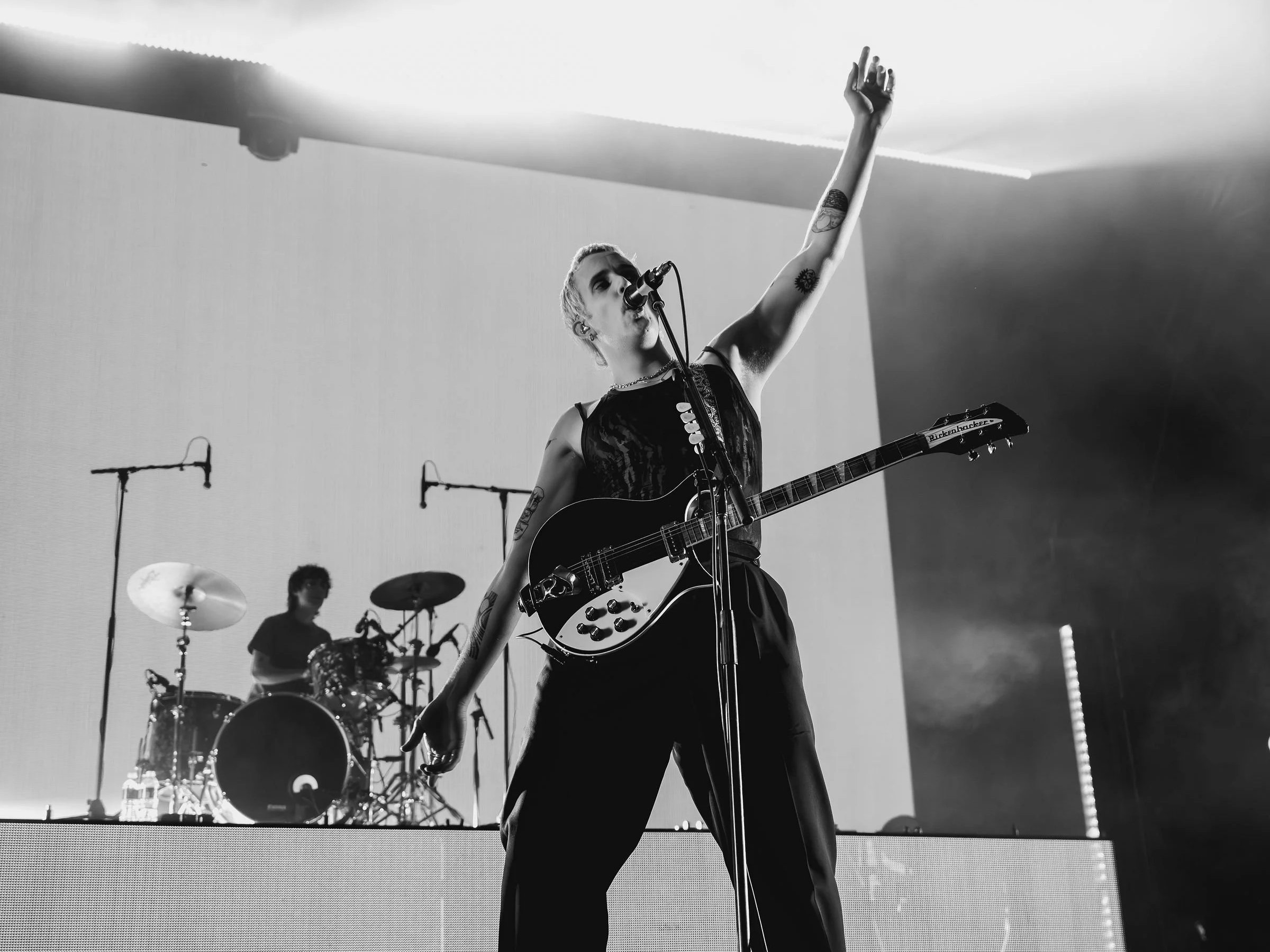 A black and white photo of a female singer with short blonde hair, tattoos on her arms, and wearing a sleeveless top, performing on stage with a guitar. She is singing into a microphone while raising her right hand. Behind her, a drummer and a keyboa
