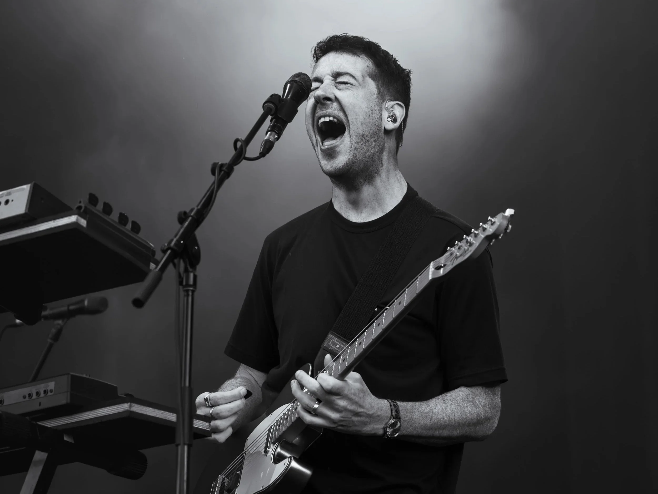 A black-and-white photo of a man singing into a microphone while playing an electric guitar, with electronic equipment nearby, on a gray background.