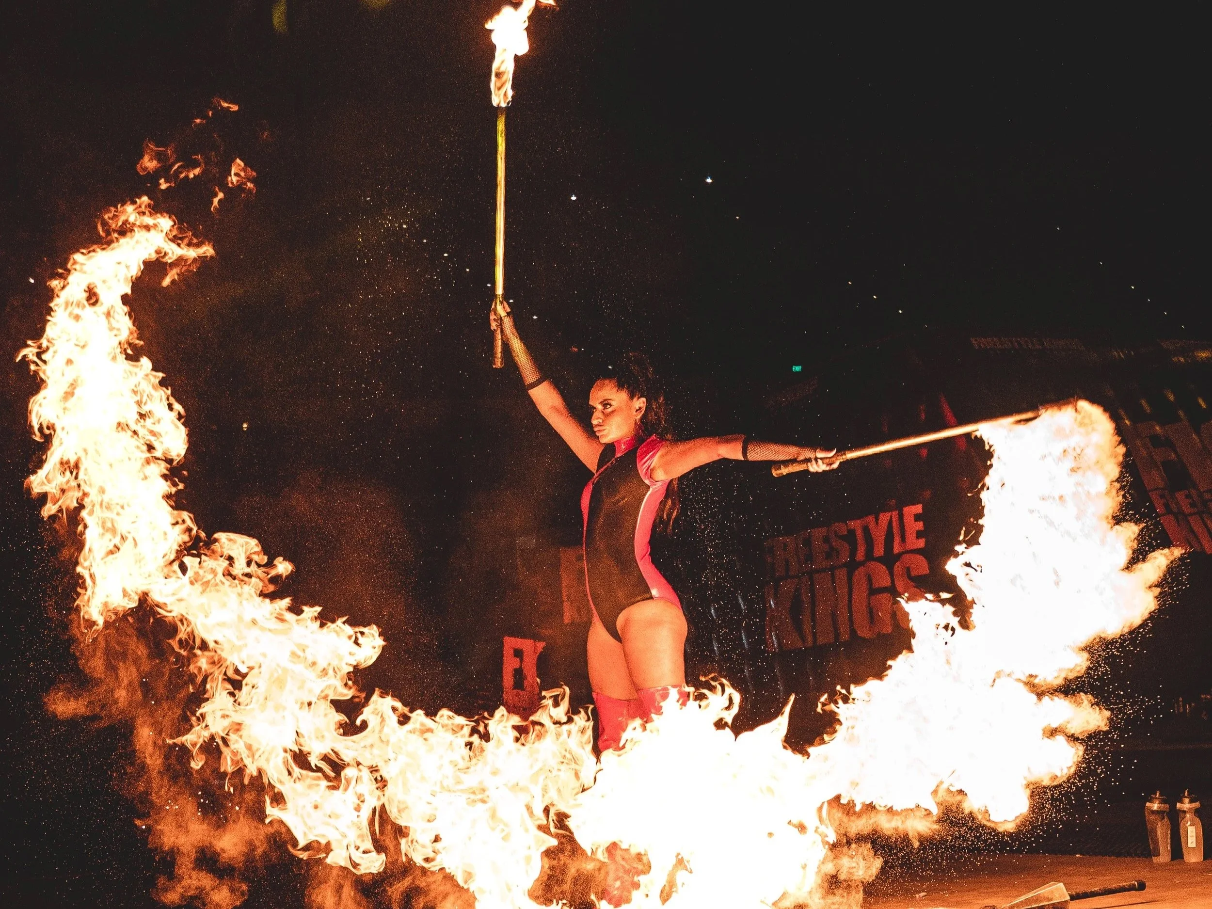 A female fire performer is balancing a fire staff above her head while standing among large flames.