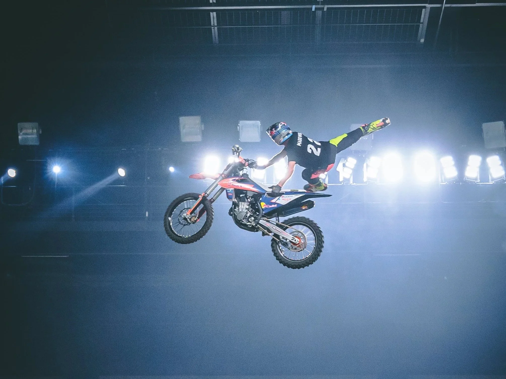 A motocross rider performing a trick in mid-air inside a dimly lit indoor arena with bright lights in the background.