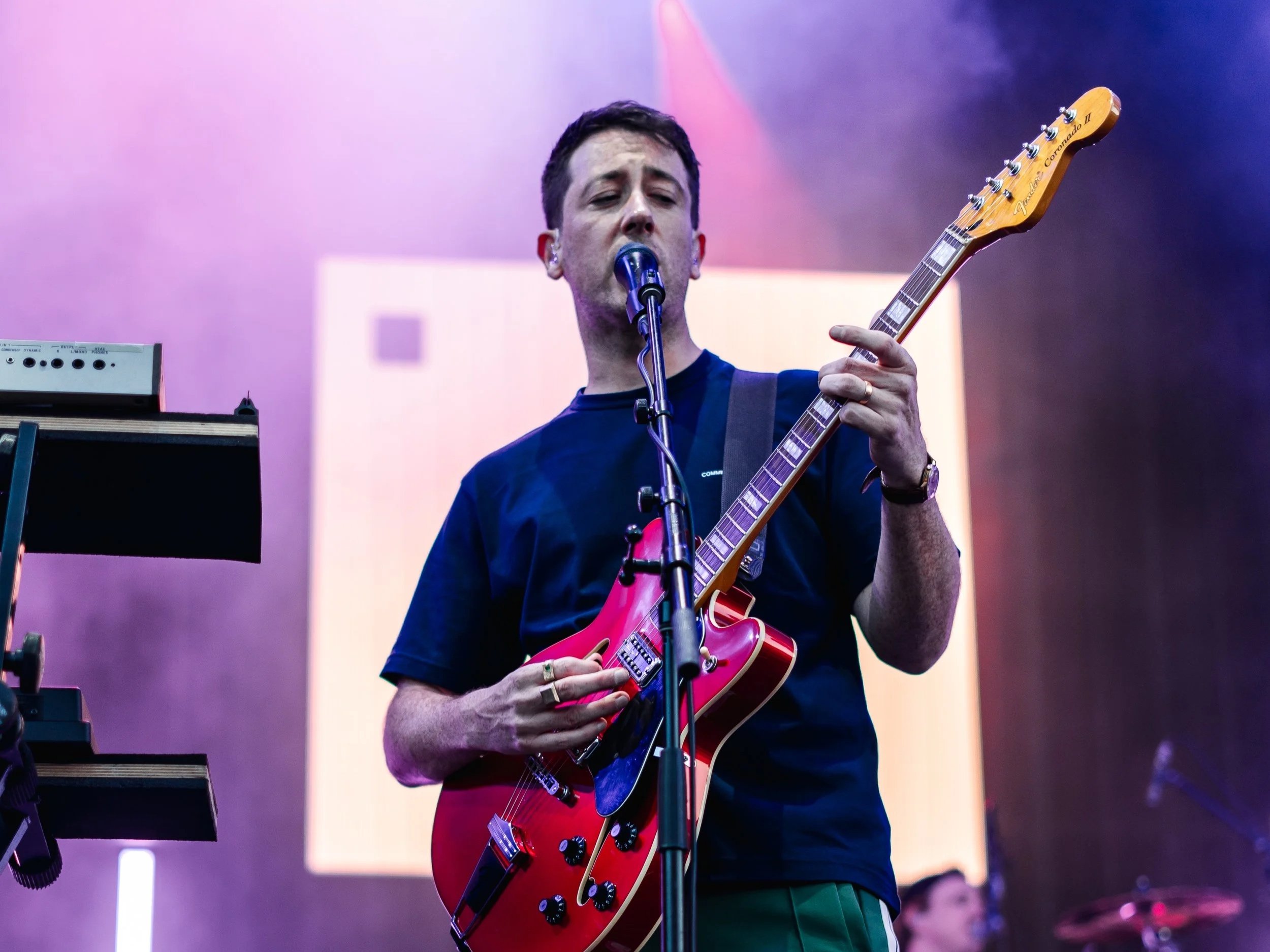 A man singing into a microphone while playing a red electric guitar on stage with purple and pink lighting.