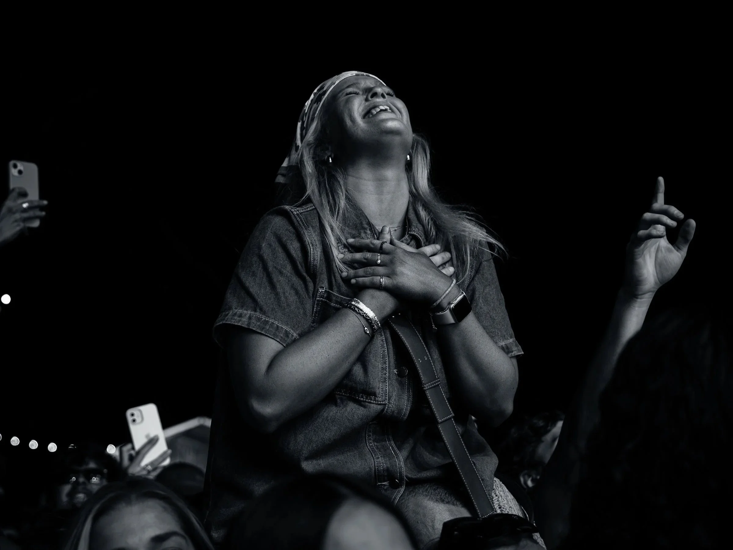A woman with long hair and a headband, dressed in denim, experiencing an emotional moment with a hand on her chest and eyes closed, surrounded by other people taking photos in a dark setting.