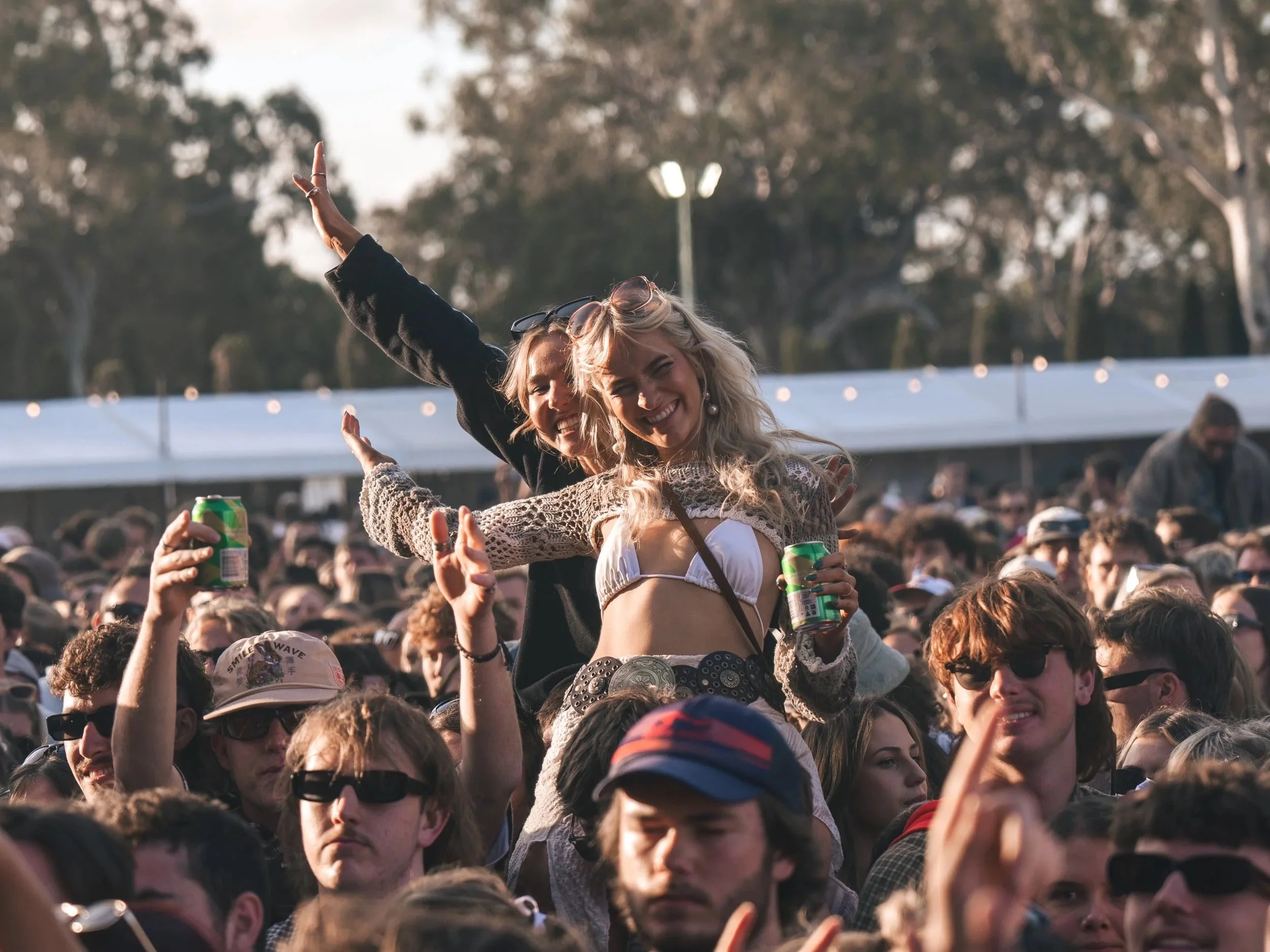 Two women are sitting on the shoulders of people in a crowded outdoor event with many attendees. Both women are smiling and holding drinks, and one is making a peace sign with her hand. The background features trees and a white tent.