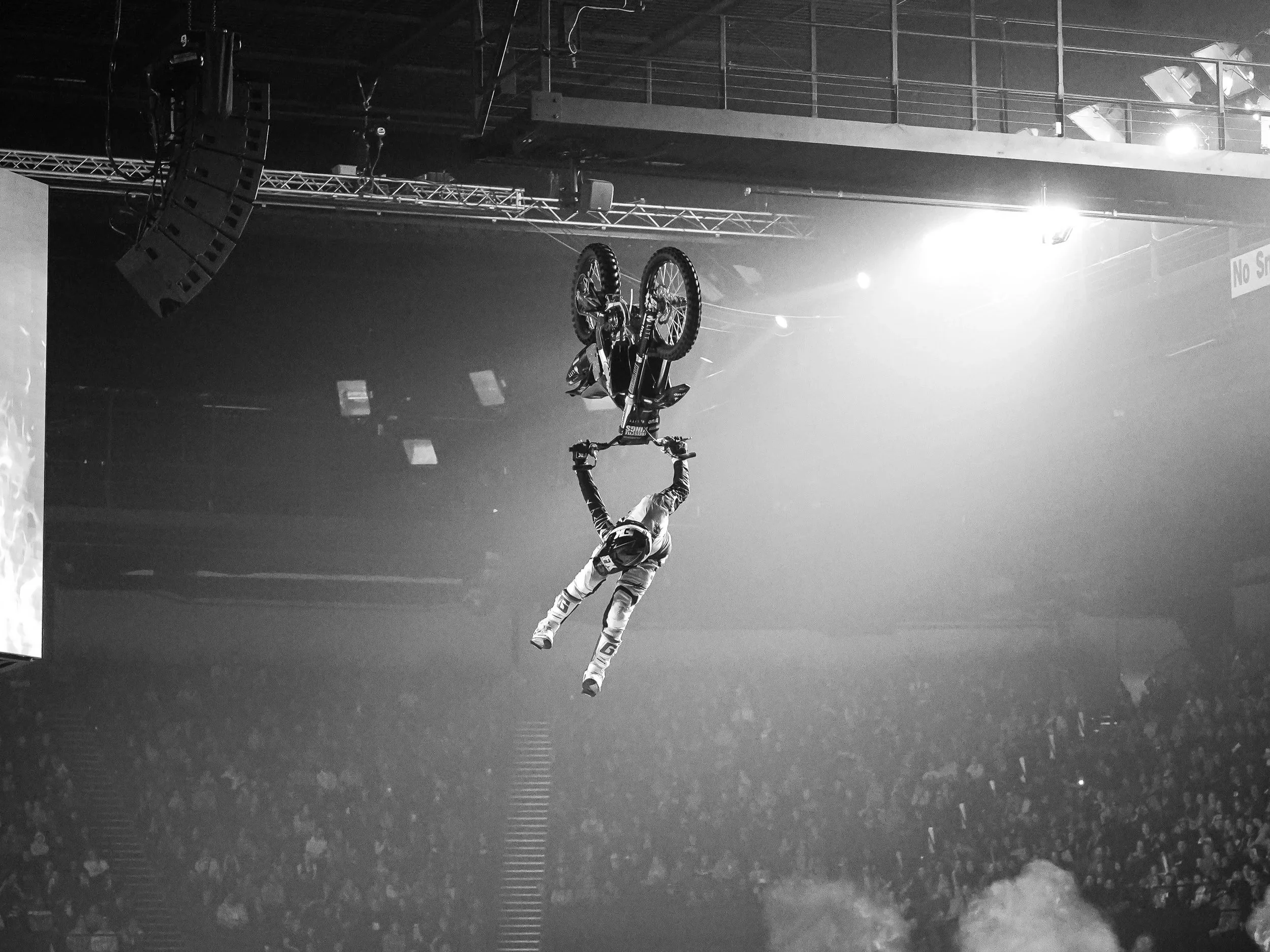 An athlete performing a stunt on a bicycle in an indoor arena with stage lighting and sound equipment, captured in black and white.