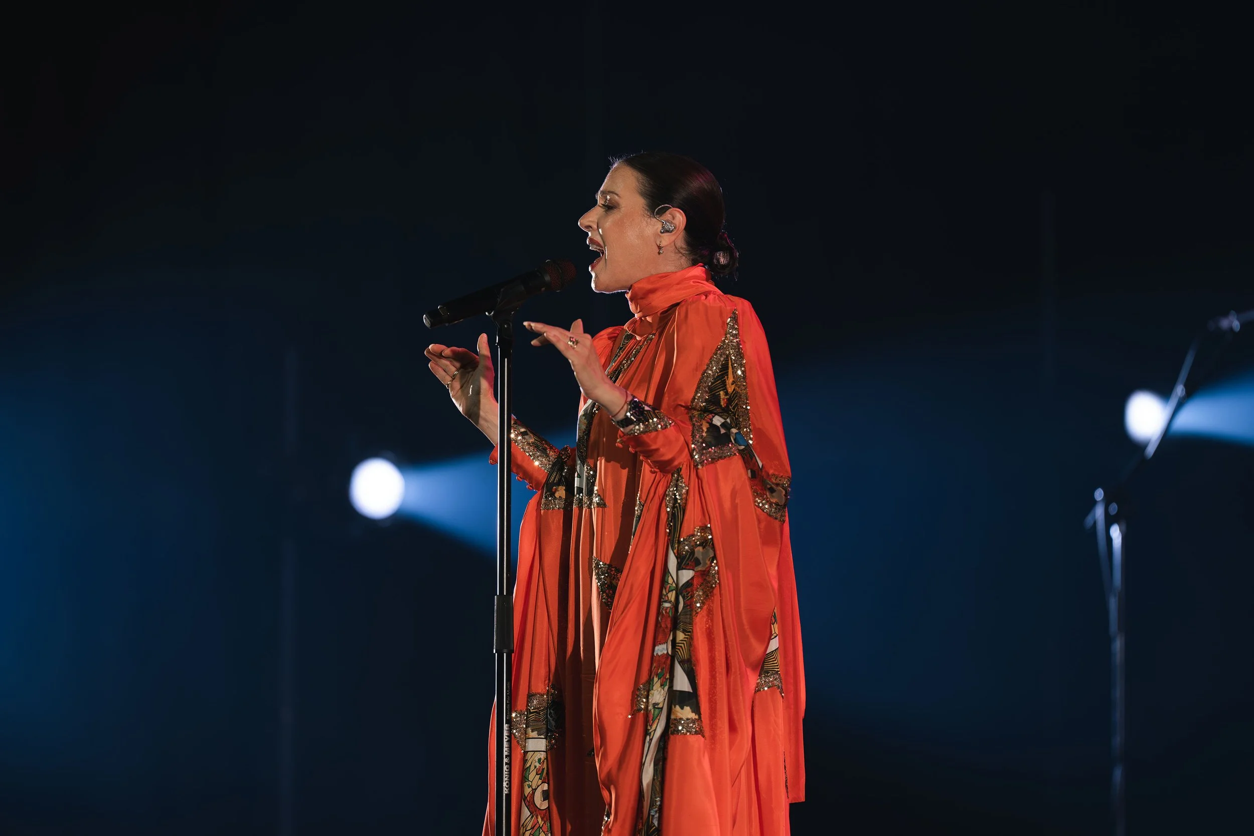 A woman singing passionately into a microphone on stage, wearing an orange and gold ornate outfit, with dark background and stage lights.