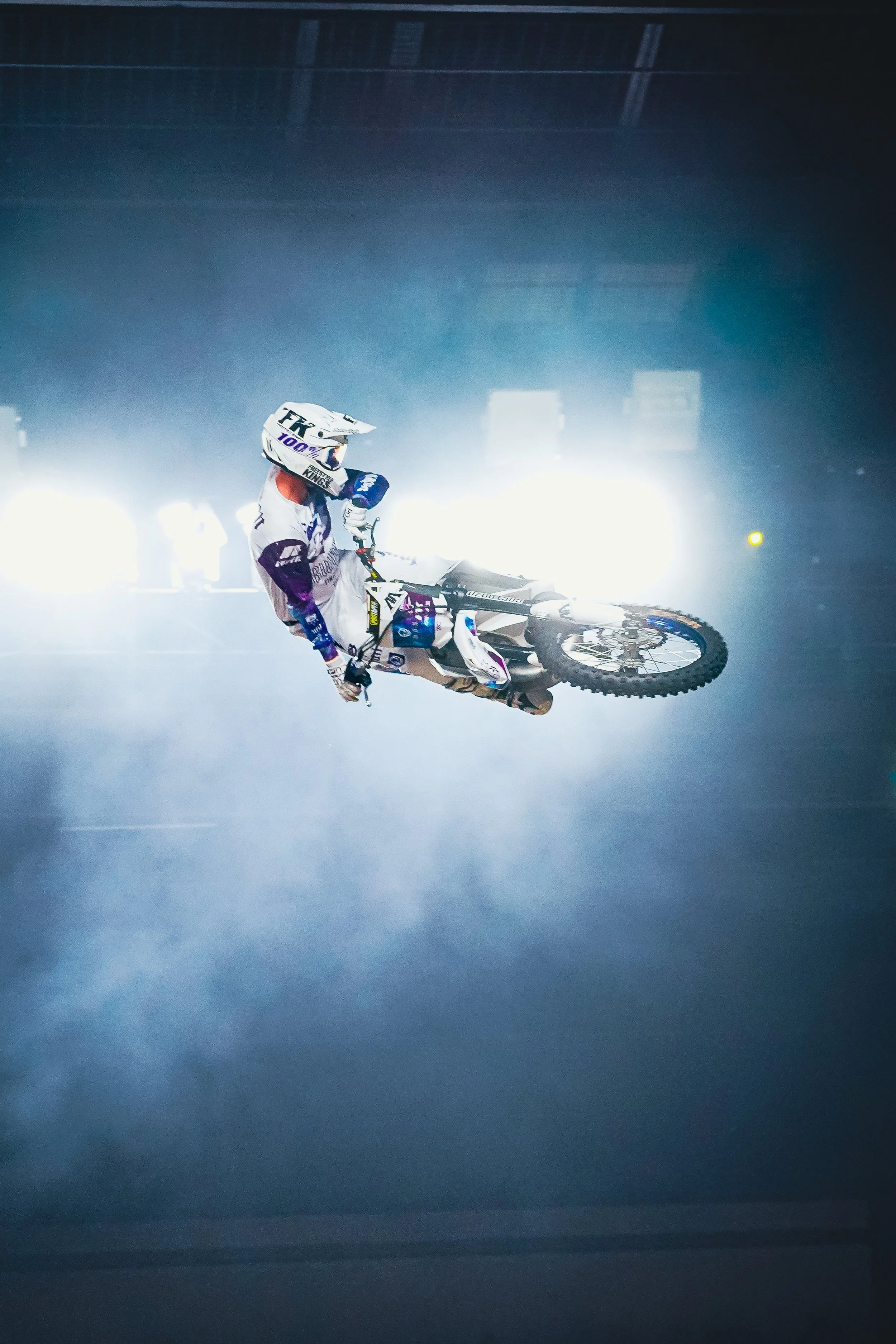 A motocross rider in white and black gear, including a helmet, riding a dirt bike, is captured mid-air against a background with bright lights and smoke, suggesting an indoor or staged stunt event.