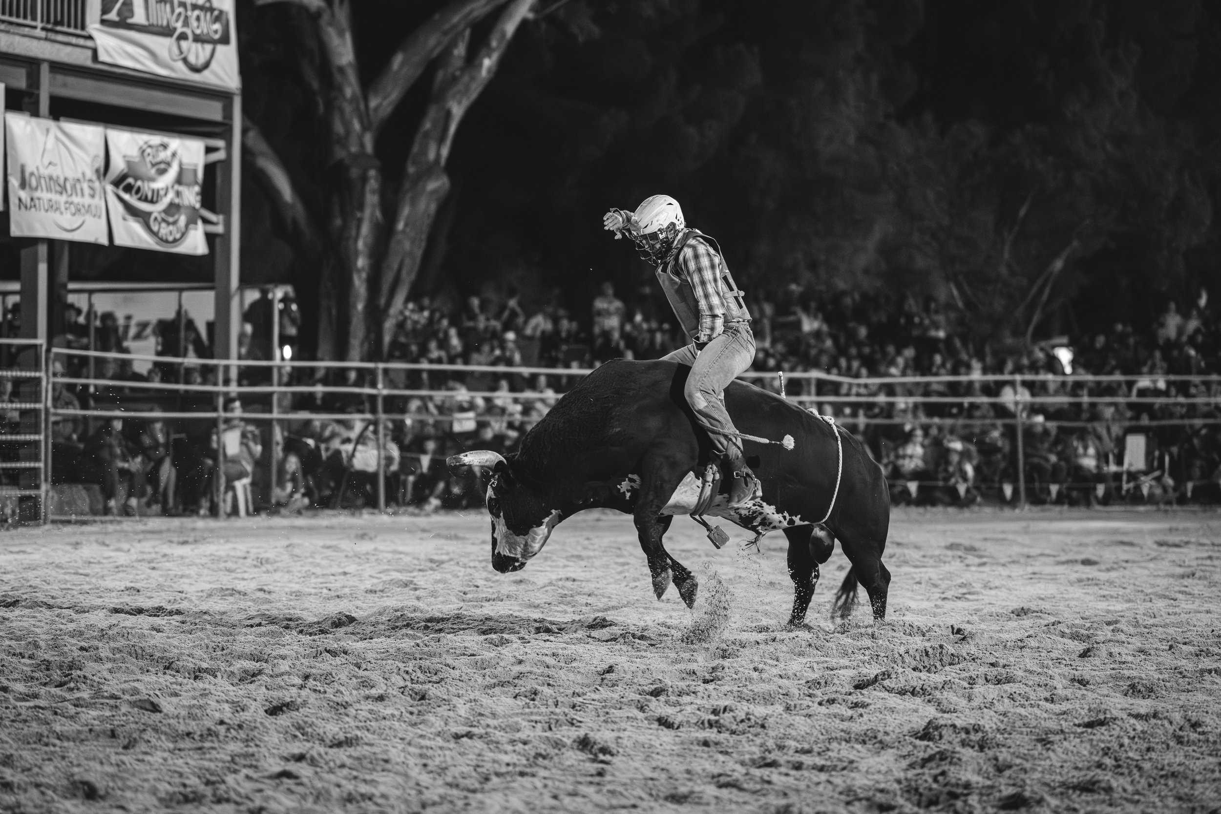 A rodeo event at night, featuring a cowboy in a helmet riding a bucking bull on a dirt arena, with an audience in the background.
