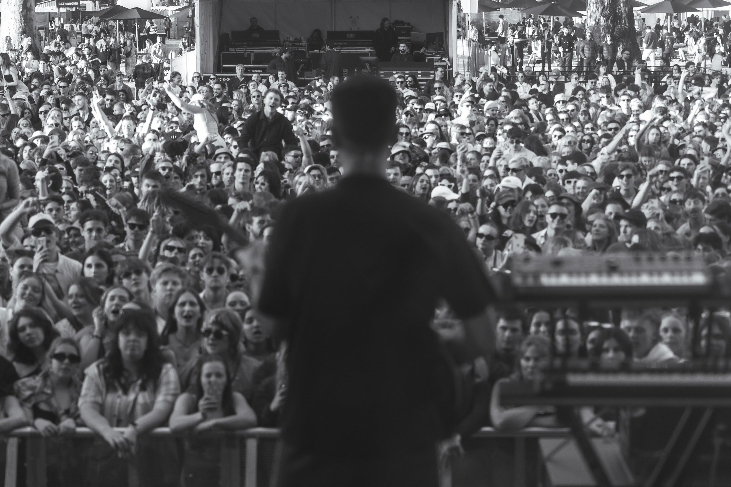 A crowd of people attending an outdoor event, viewed from behind a person standing at a DJ booth, with a stage or booth with equipment and DJs in the background.