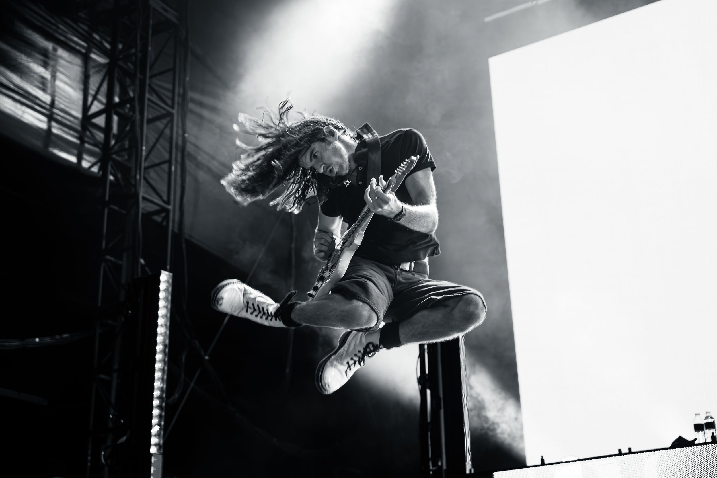 A male guitarist performing on stage, jumping in the air with his hair flying, playing an electric guitar, with stage lights and scaffolding in the background.